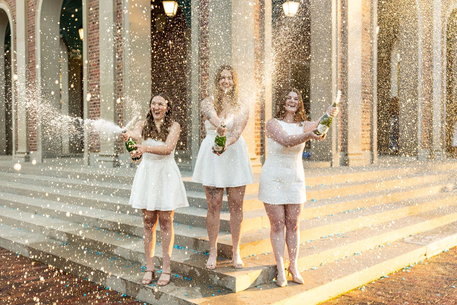 Three women in white dresses celebrate with champagne spray on elegant building steps.