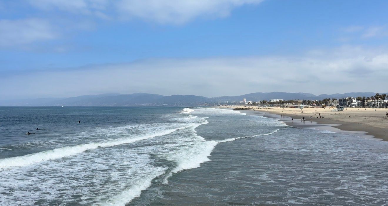Aerial view of Venice Beach coastline with rolling waves, sandy shore, and Santa Monica mountains in the background.