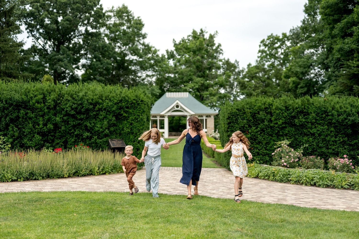 A woman and three children run joyfully through a lush garden path toward a charming gazebo surrounded by greenery.