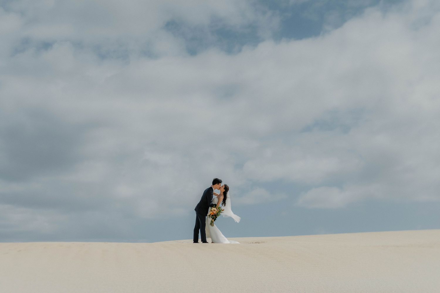 A couple embraces on a white sand dune against a cloudy blue sky in a romantic desert setting.