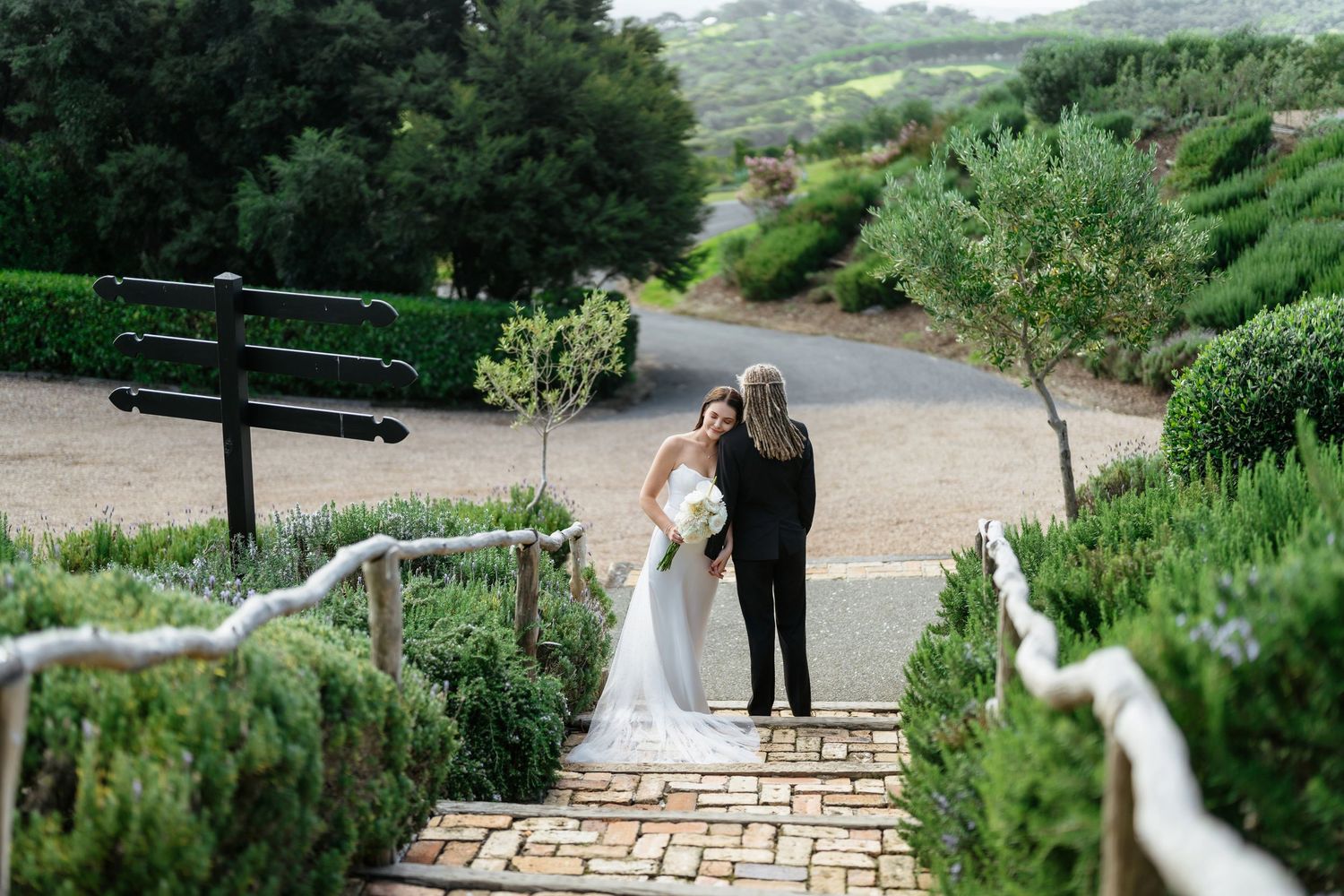 Bride in white gown sits on golden tufted sofa holding bouquet while assistant arranges veil in elegant venue.