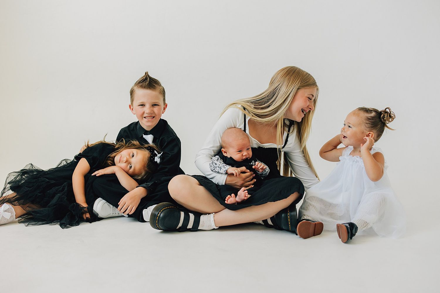 A mother sits on a white studio floor with her four children, including a newborn, dressed in black and white outfits.