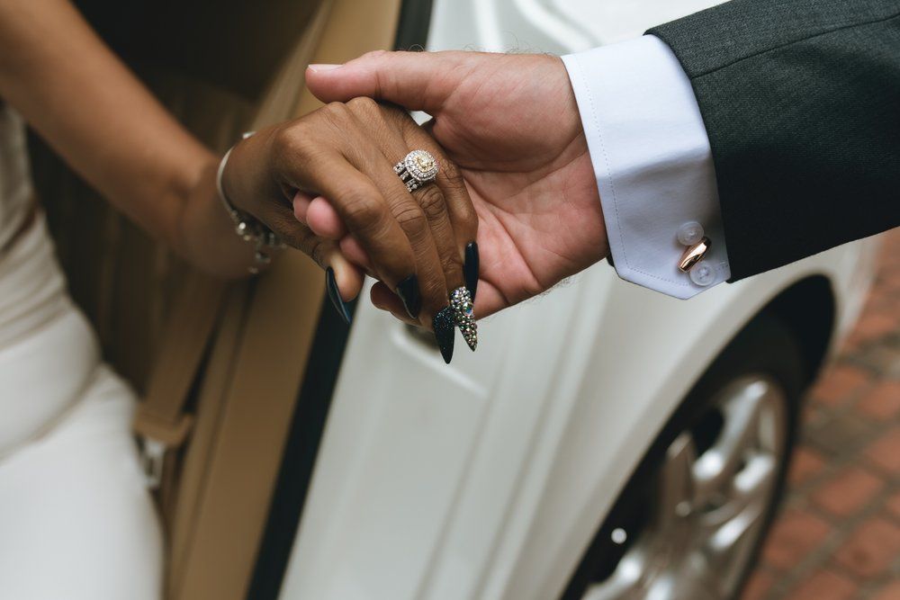 Bride and groom holding hands near a white car, showcasing an elegant diamond engagement ring and wedding bands.
