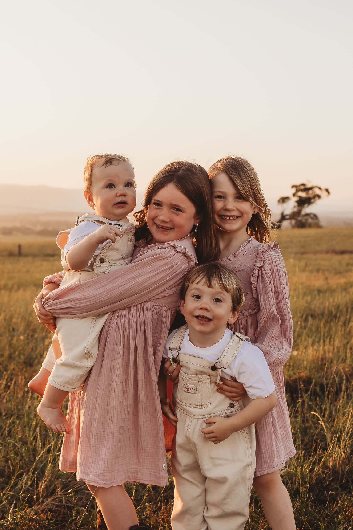 Children play and embrace in a golden field at sunset wearing pink dresses and neutral colored outfits.