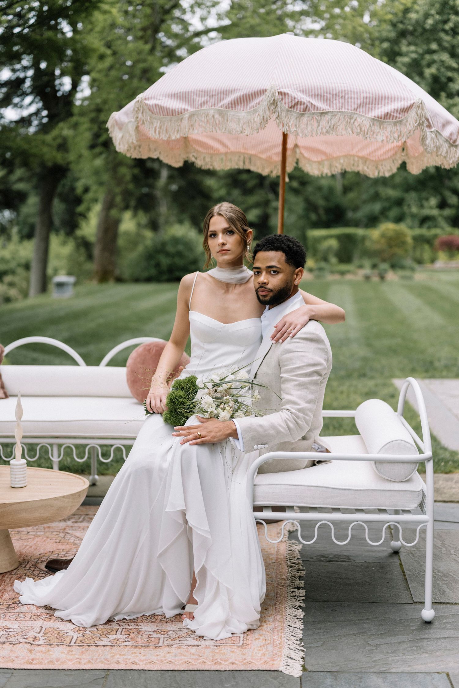 Bride and groom sit together on white outdoor furniture under a pink fringed umbrella in a lush garden setting.