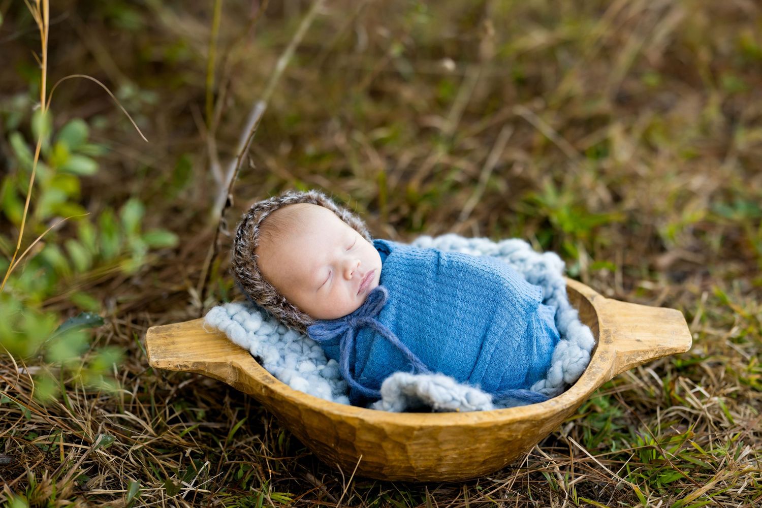 Sleeping newborn wrapped in blue swaddle with knit bonnet resting in wooden bowl on grass.