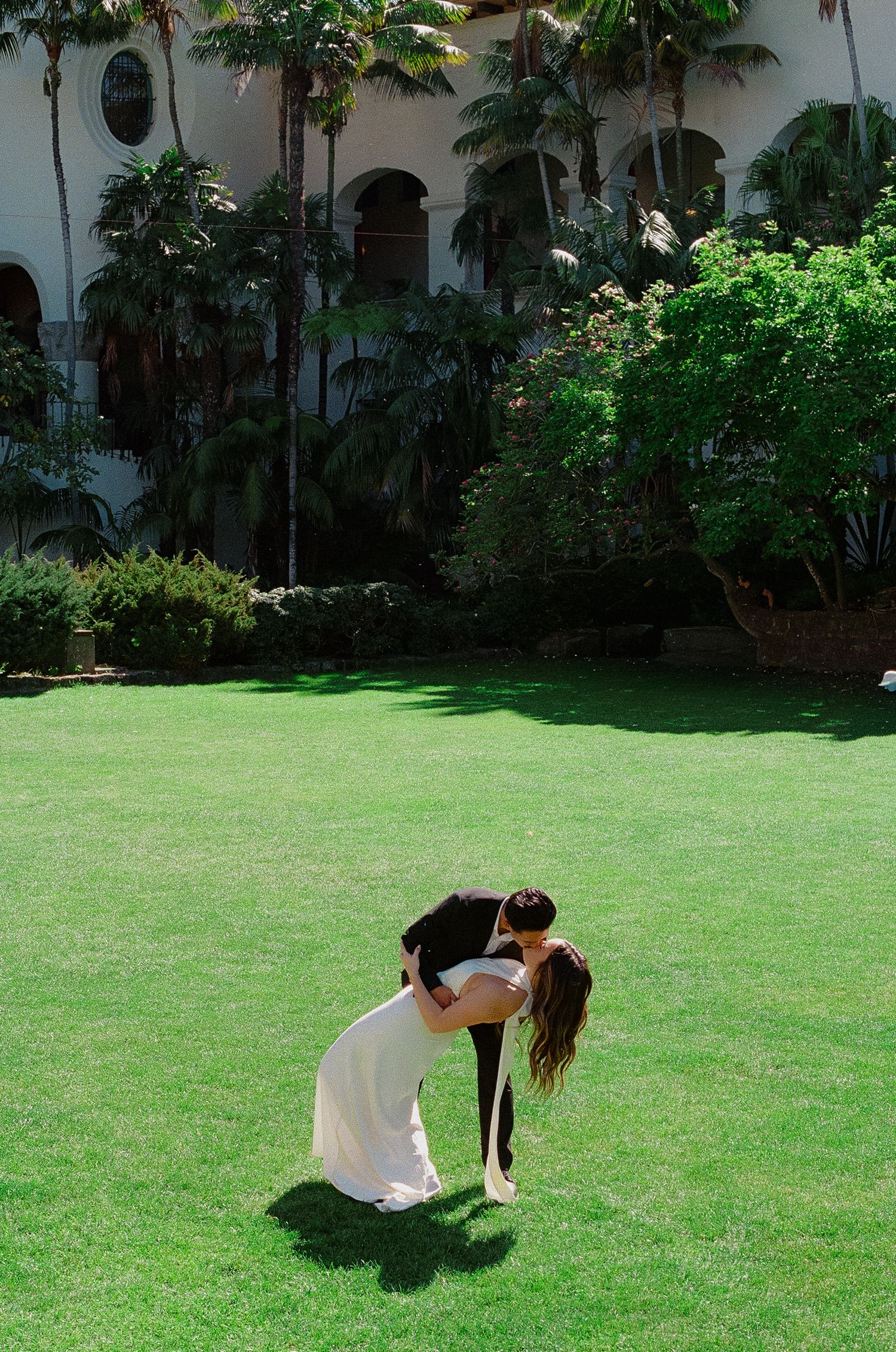 A couple shares a romantic dip kiss on a lush green lawn with palm trees and a white building backdrop.
