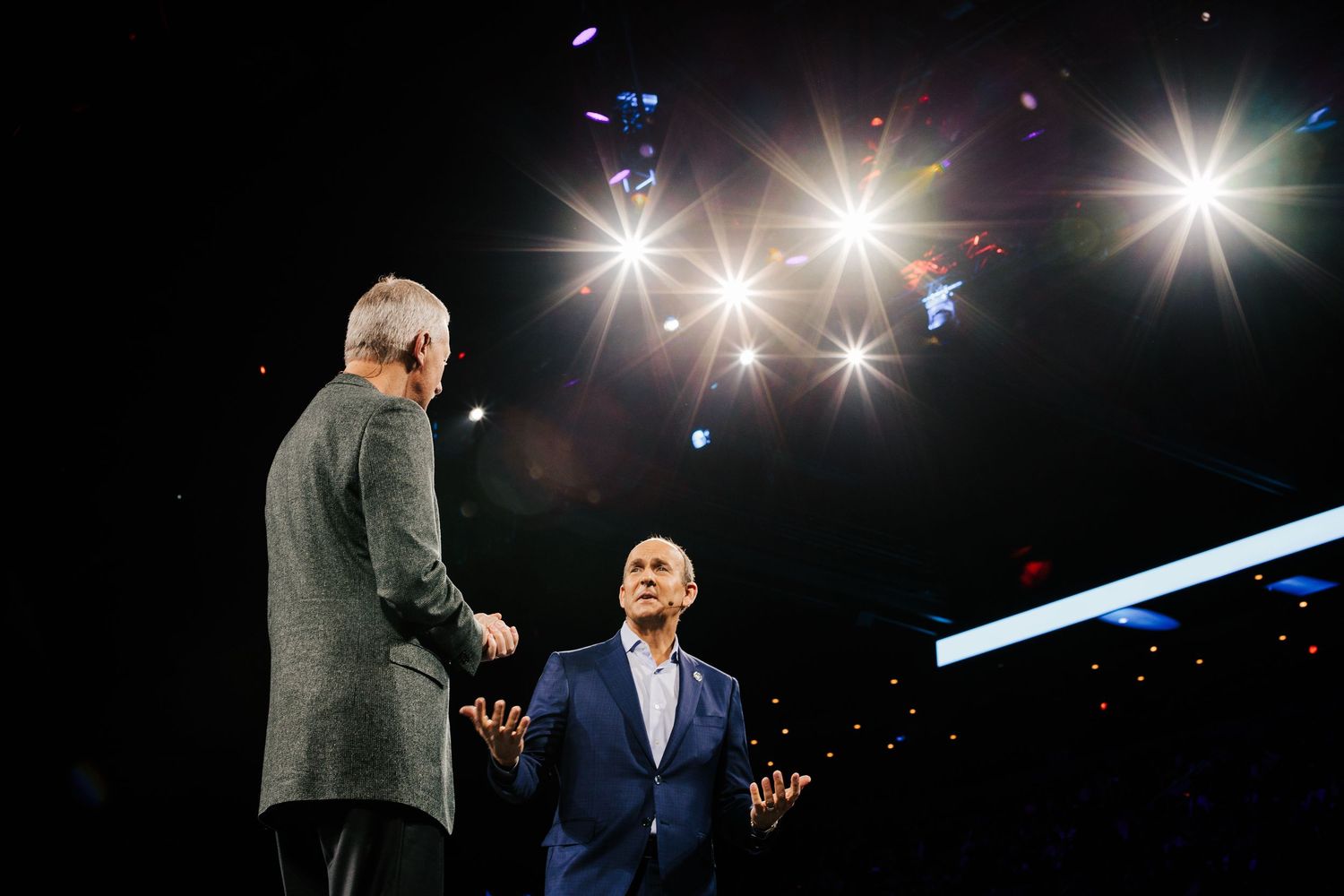 Two people in suits interact on a dark stage with bright starburst lights overhead during an event.