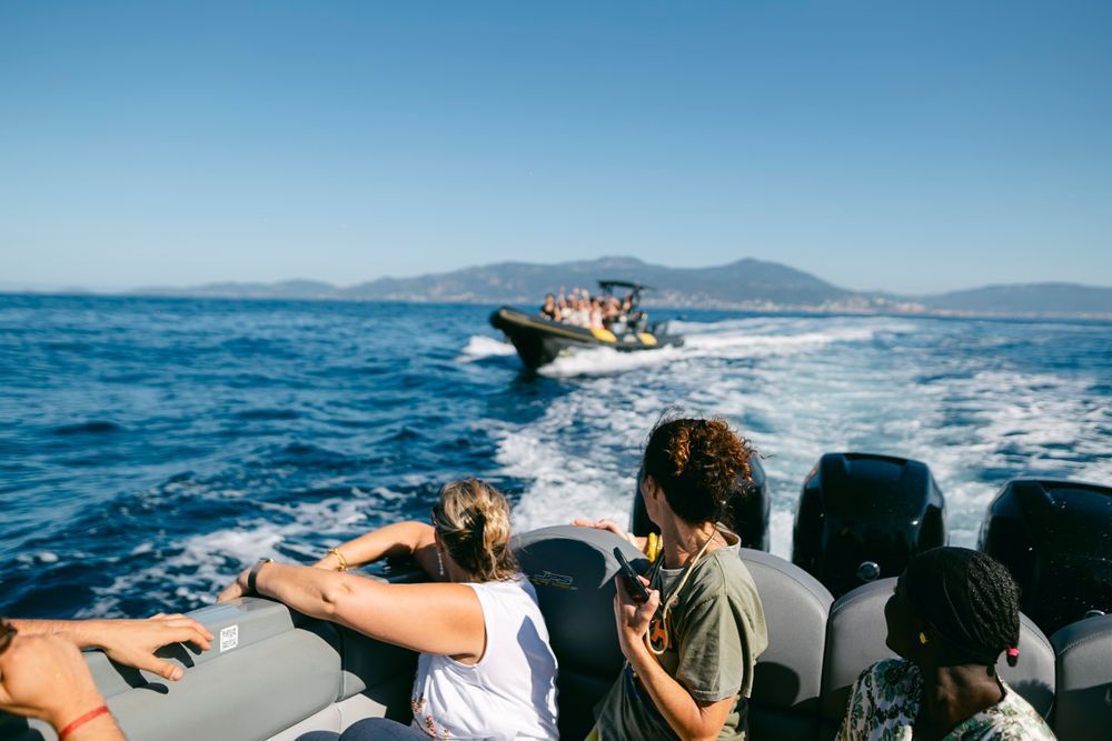 People on a speedboat watching another boat race across blue ocean waters with mountains in the background on a sunny day.