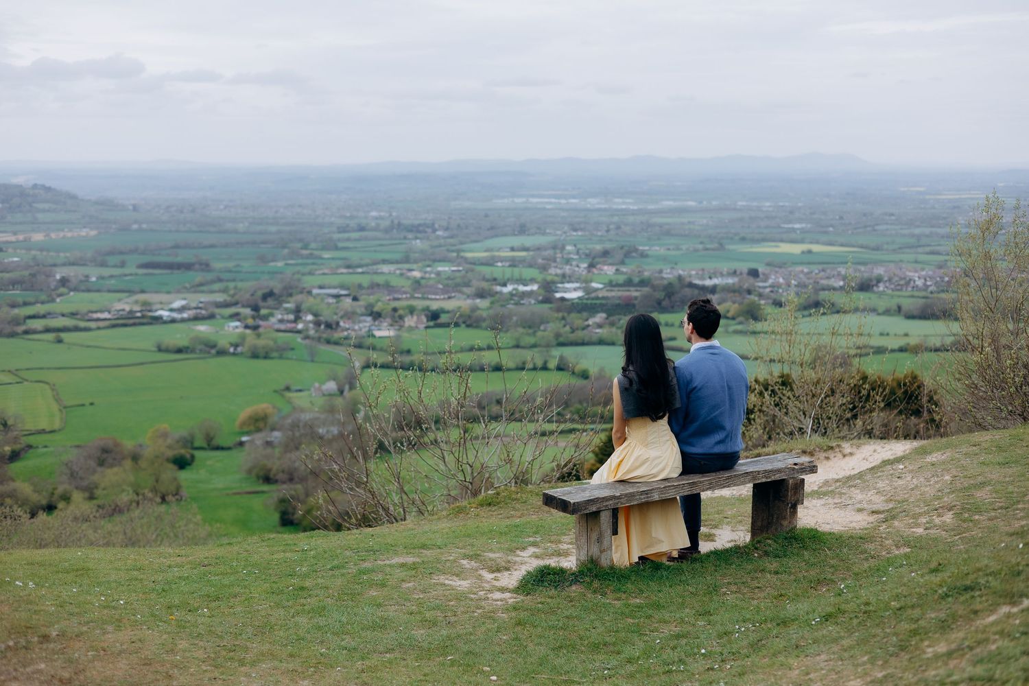 Couple sitting on a wooden bench overlooking a scenic green valley landscape from a hilltop viewpoint.