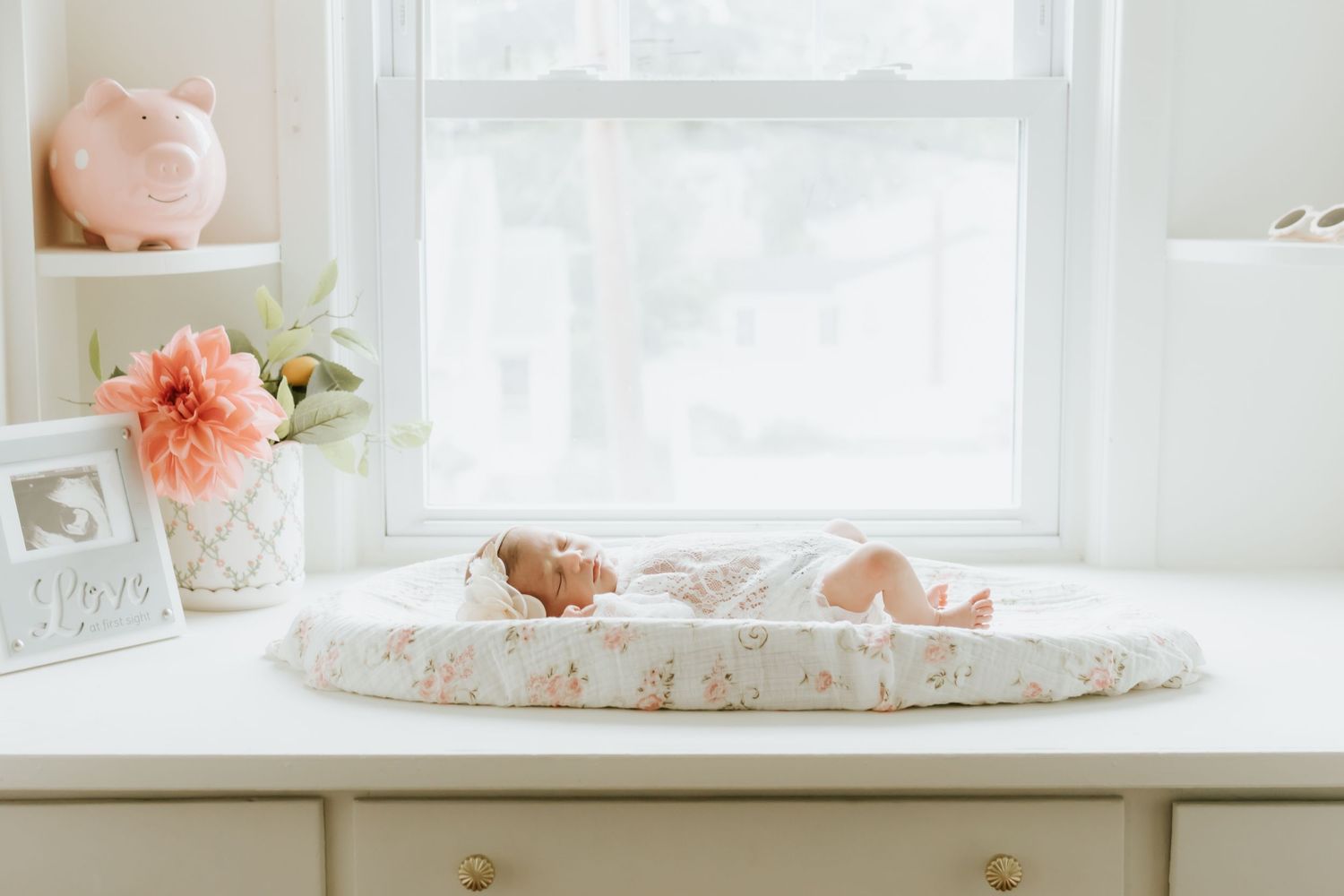 A serene white nursery with a newborn on a changing table near a bright window, teddy bear, and soft pink decor. nj newborn photographer