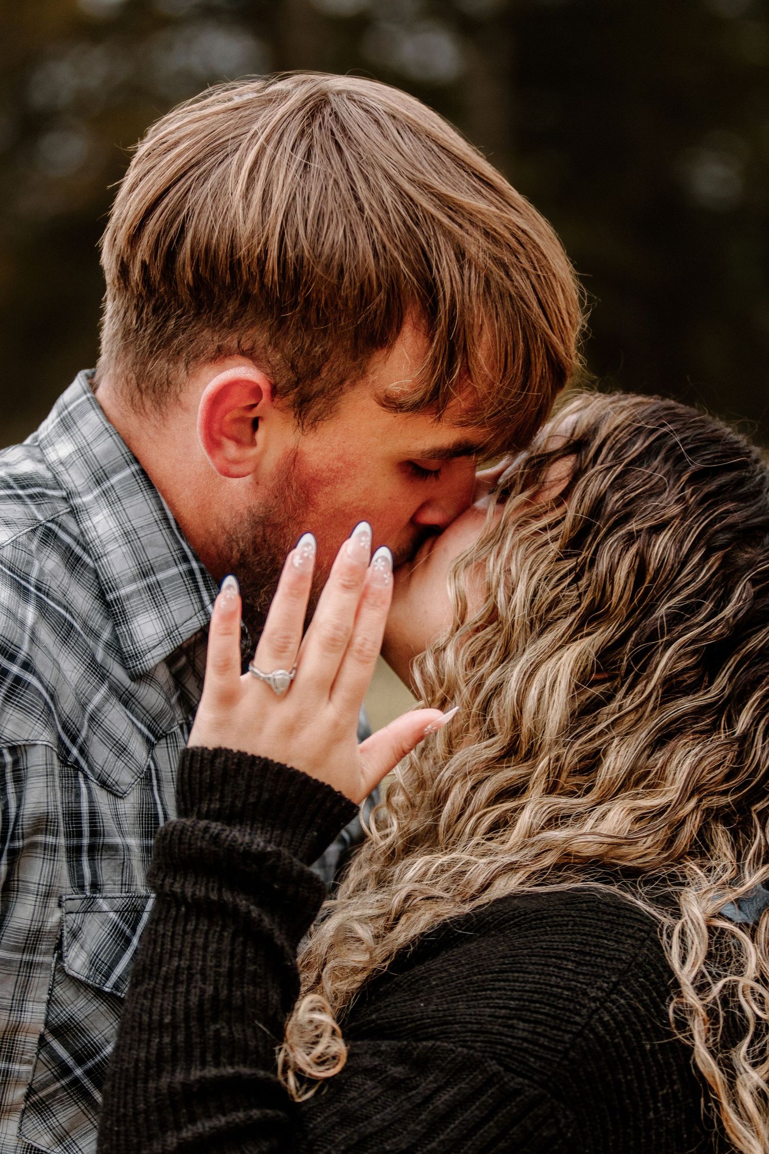 An engagement ring sparkles as a couple shares a romantic kiss outdoors.