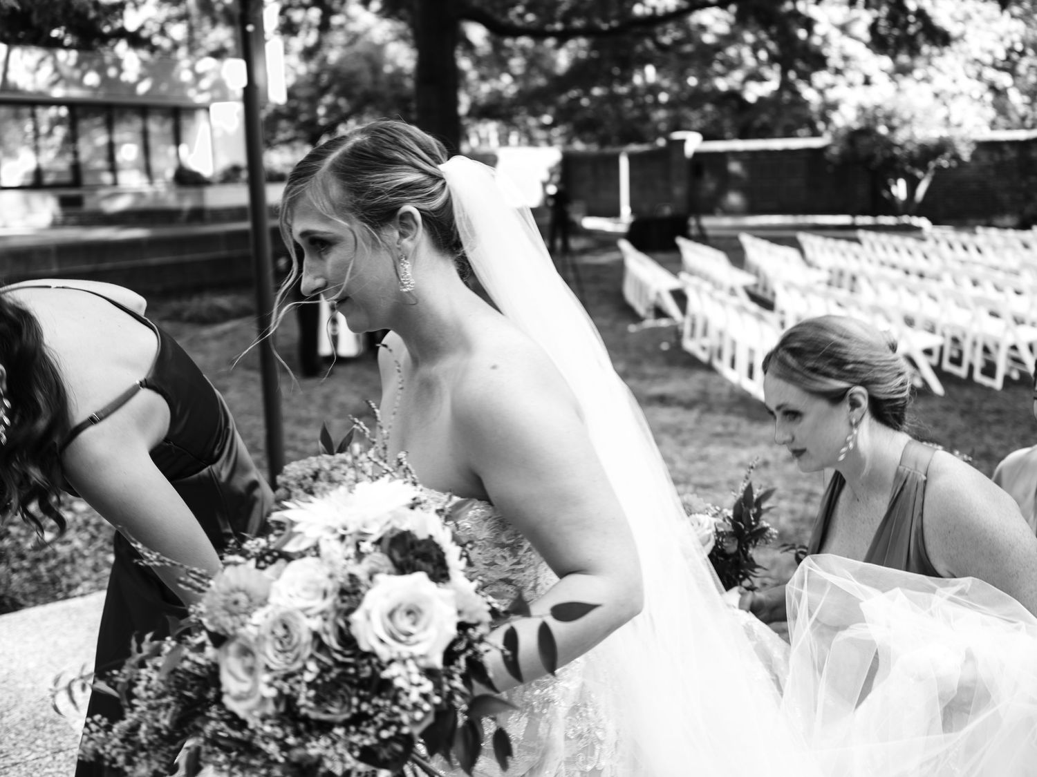 Black and white photo of bridal party making adjustments to wedding dress outdoors.