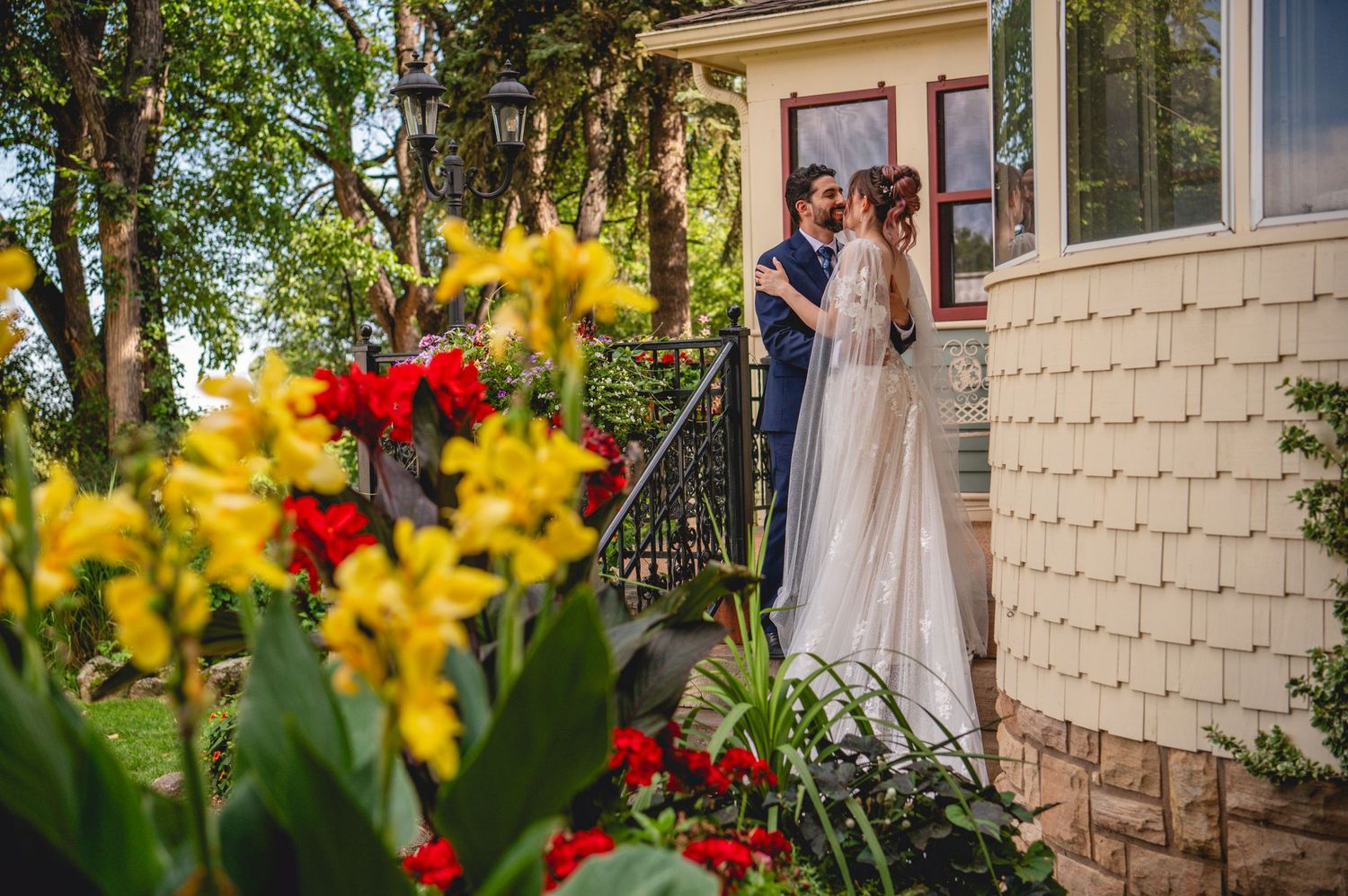 A couple stands on the front porch steps of a tan house with vibrant red and yellow flowers blooming in the foreground.