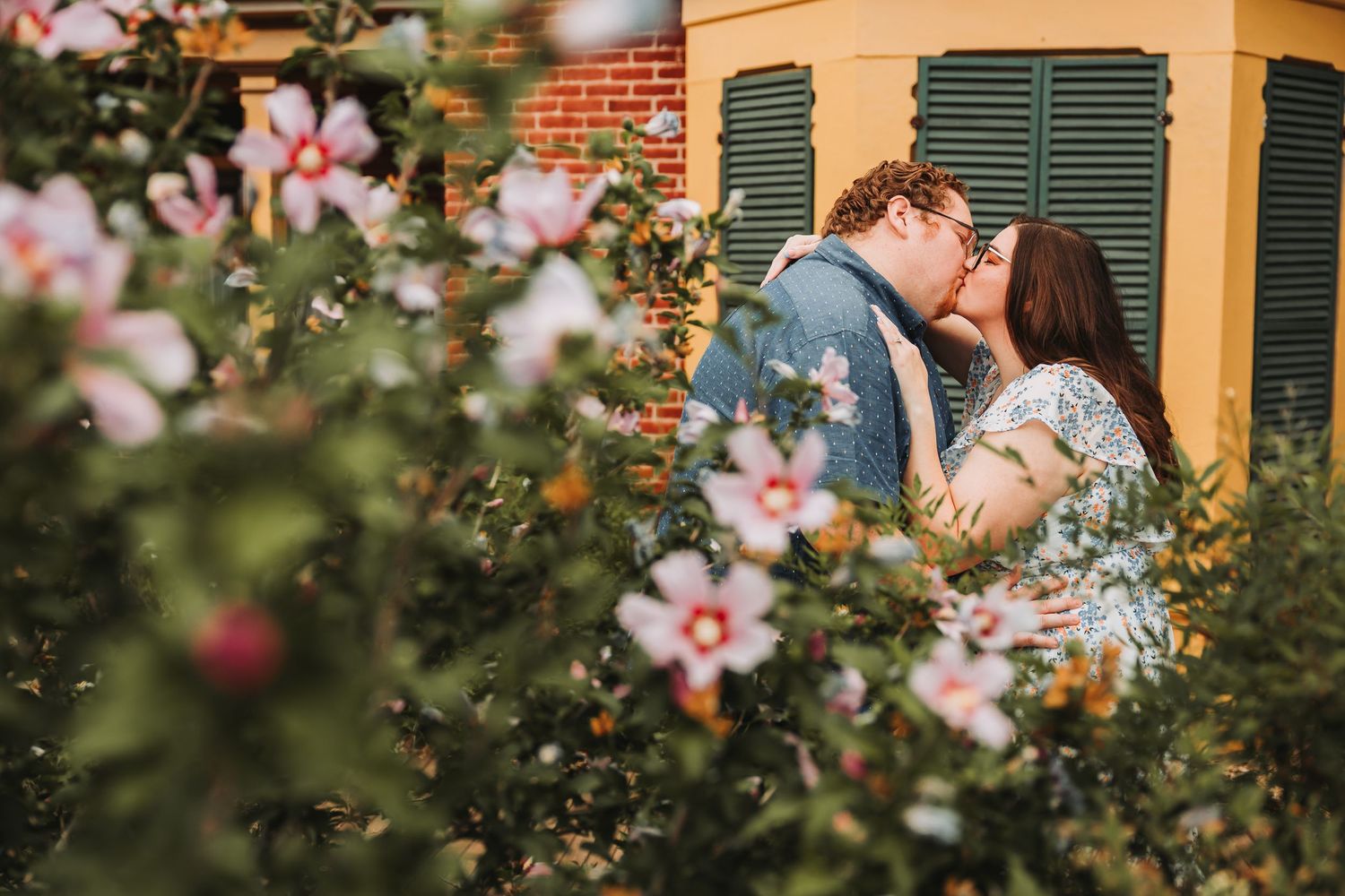 A couple shares a romantic kiss in a garden surrounded by blooming pink flowers and green foliage.