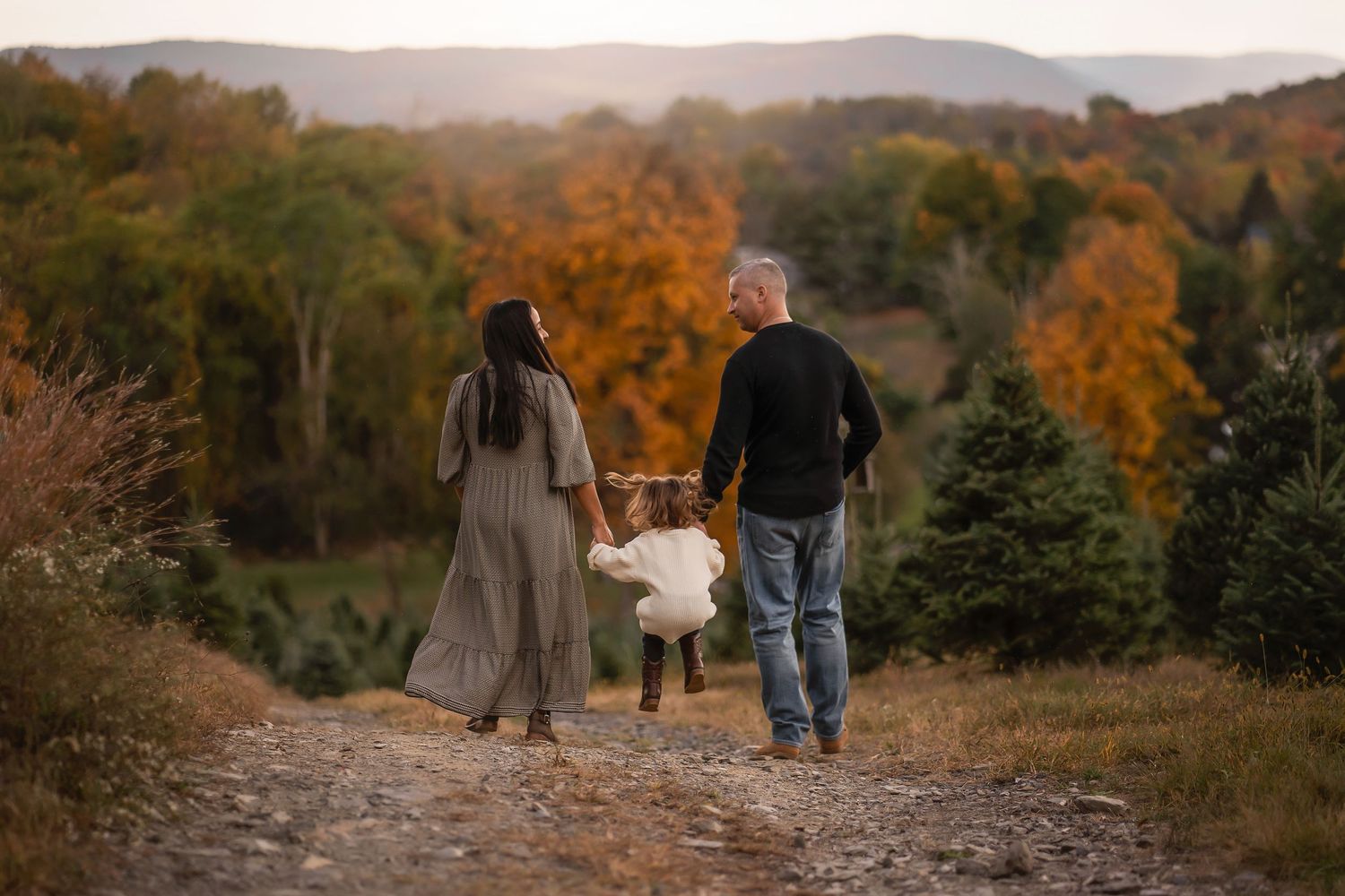 A couple walks with a child on an autumn mountain trail surrounded by golden trees at sunset.
