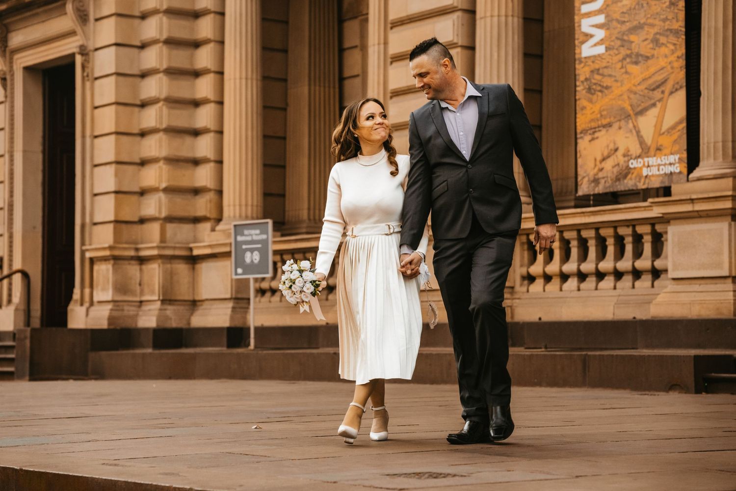 A couple in wedding attire walks together on ornate stone steps of a historic building with classical architecture.