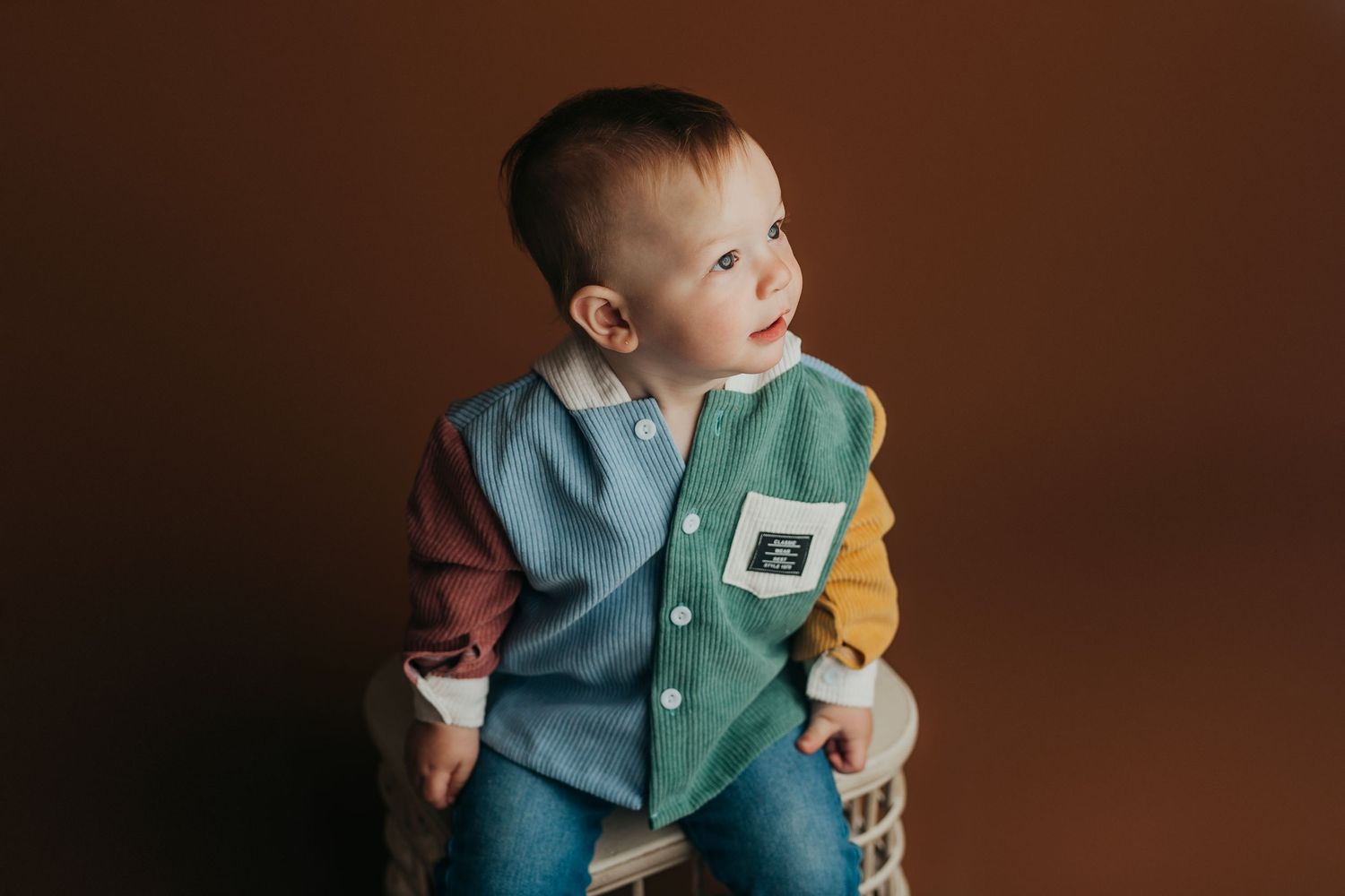 Young child in colorblock button-down shirt sits on woven stool against brown background.