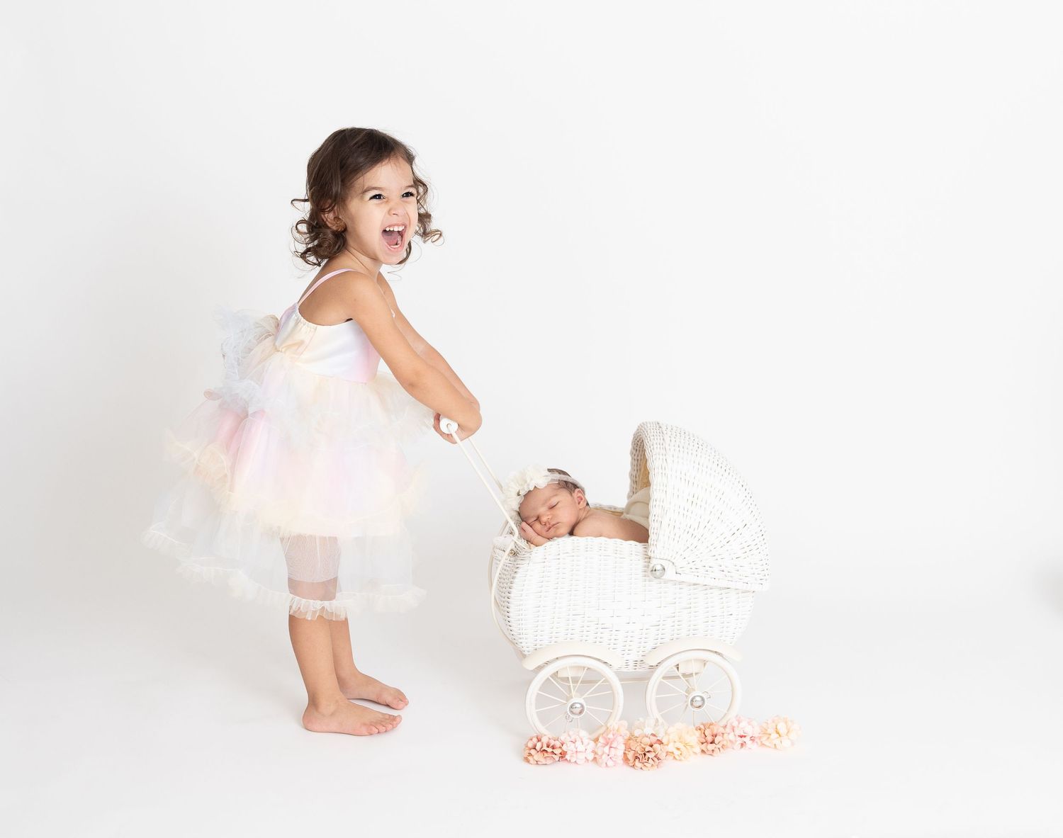 A toddler in a tutu dress excitedly peaks into a white vintage pram while standing barefoot in a bright studio setting.