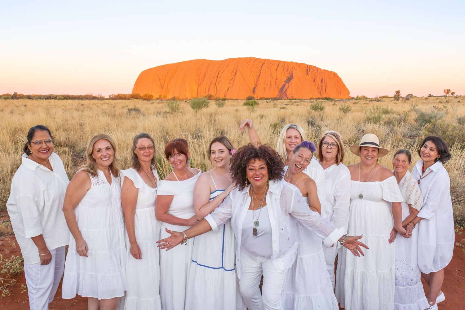 A group in white outfits stands together in front of Uluru rock formation in Australia at sunset.