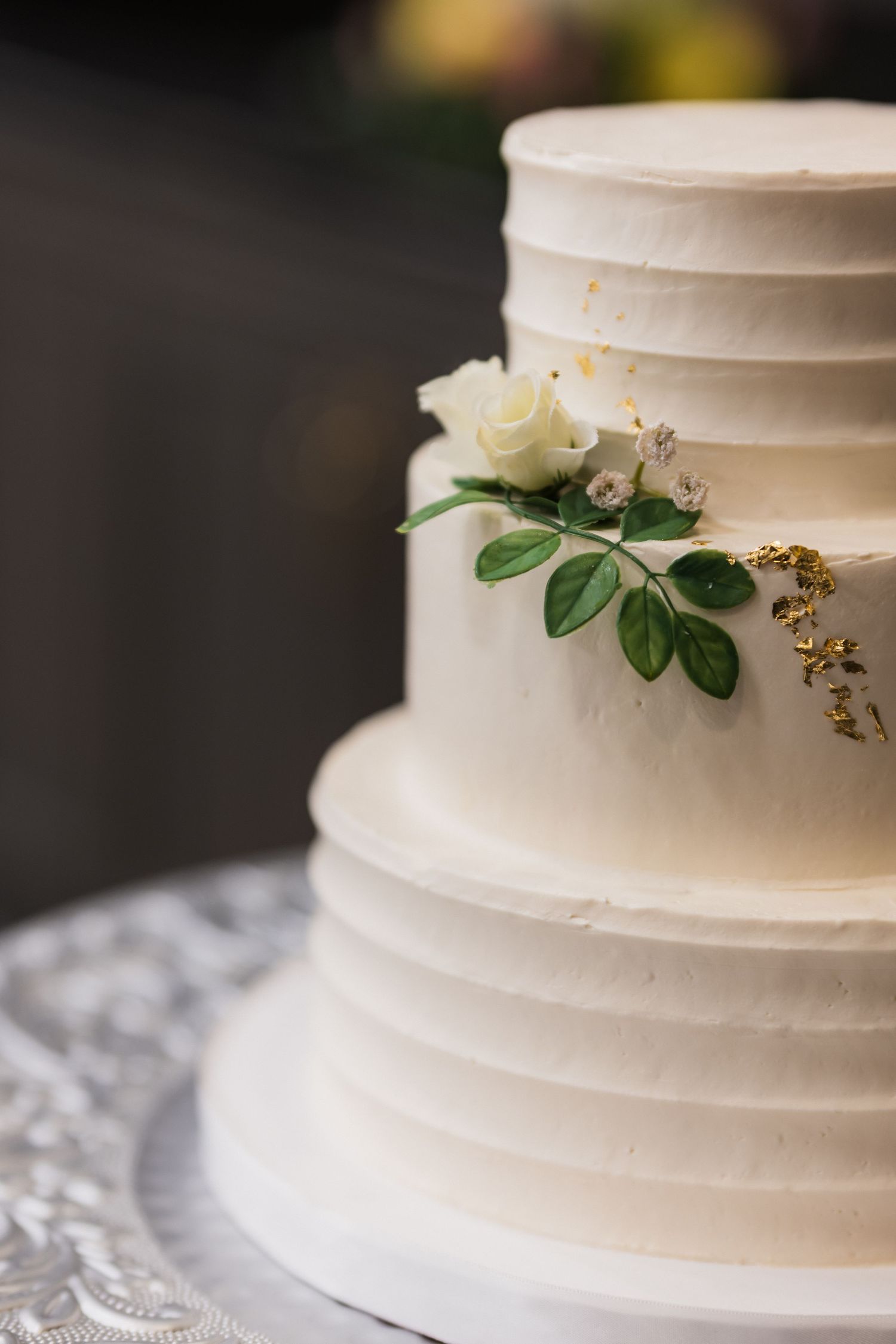 Elegant white tiered wedding cake with delicate horizontal lines and simple floral accents with green leaves on lace tablecloth.