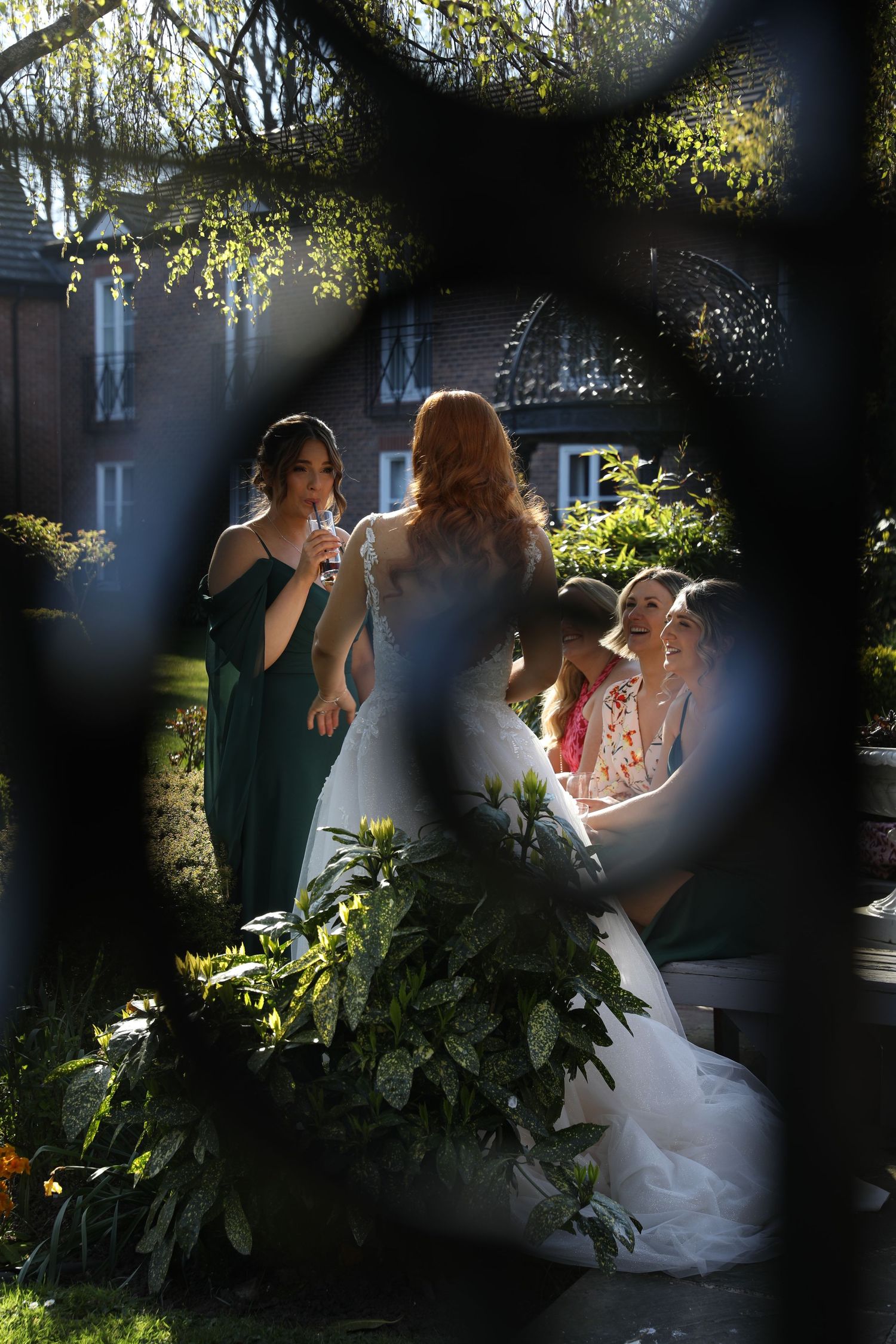 Bridal party gathers outdoors in a garden setting with green foliage and filtered sunlight creating a dreamy atmosphere.
