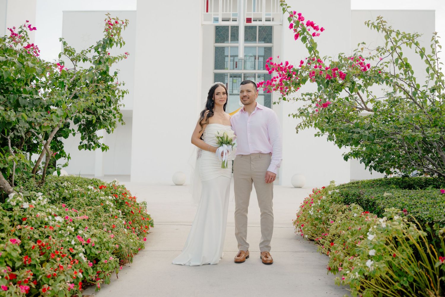 A romantic garden walkway with pink and red flowers blooming alongside a white stucco wall in bright sunlight.