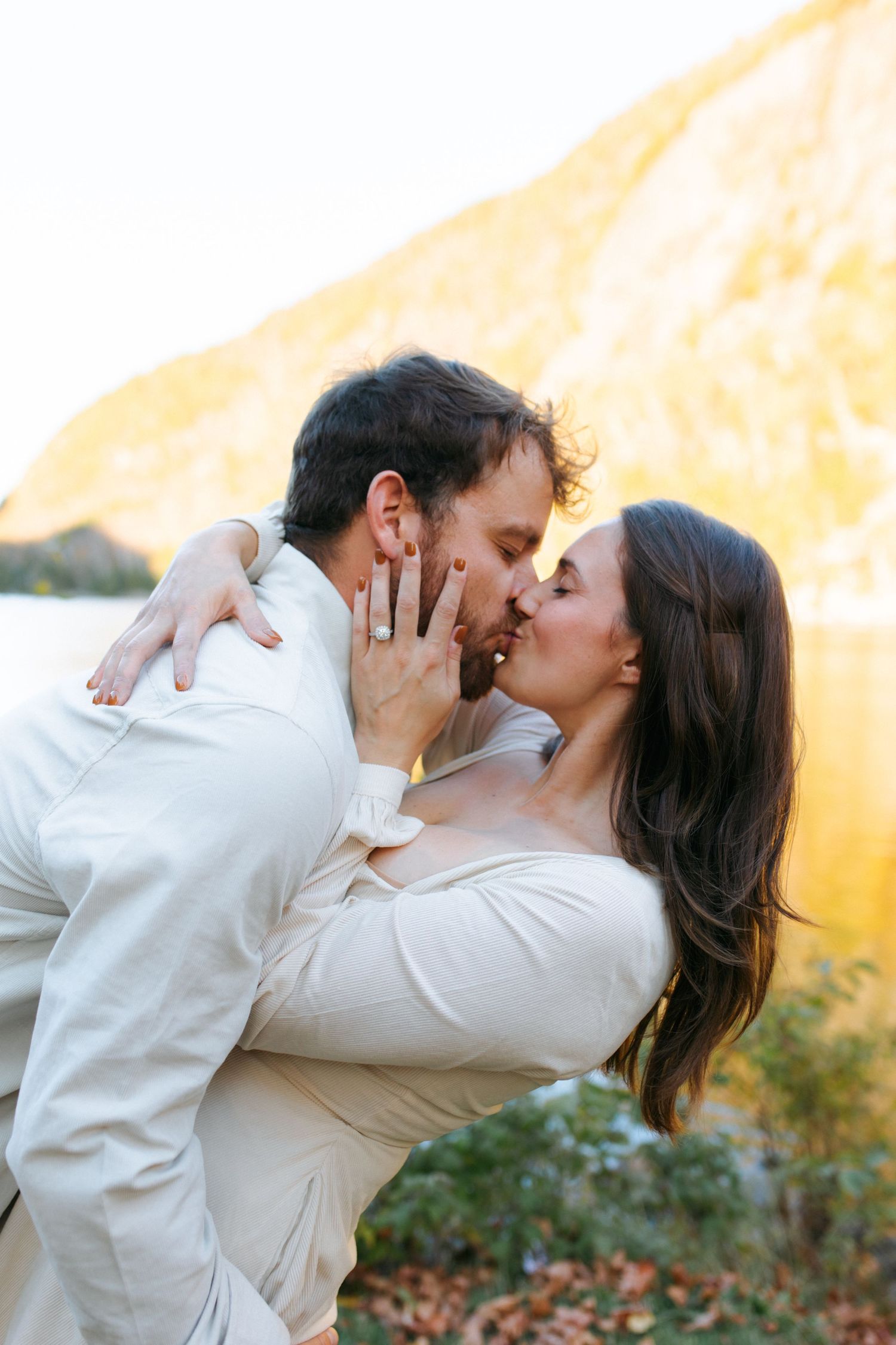 A romantic couple in cream colored sweaters share tender kisses against a warm sunset backdrop in autumn.