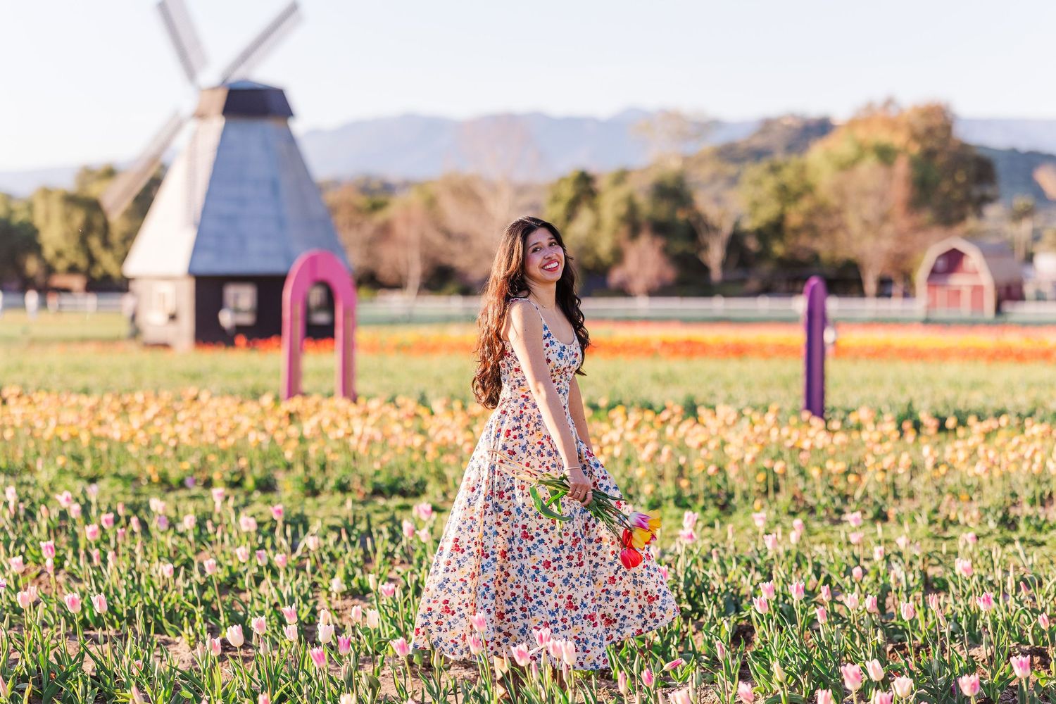 A person in a floral dress walks through a vibrant wildflower field during golden hour with mountains in the distance.