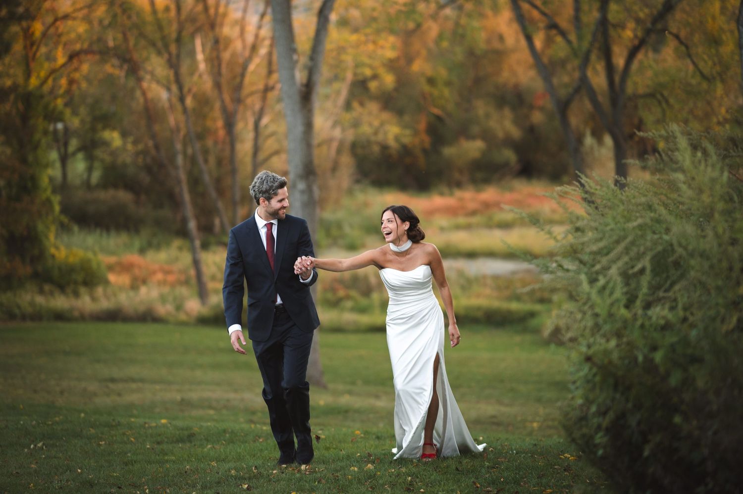 Hudson Valley wedding couple laugh and walk up hillside with fall foliage and pond behind them at Drowned Lands Brewery in Warwick, NY.