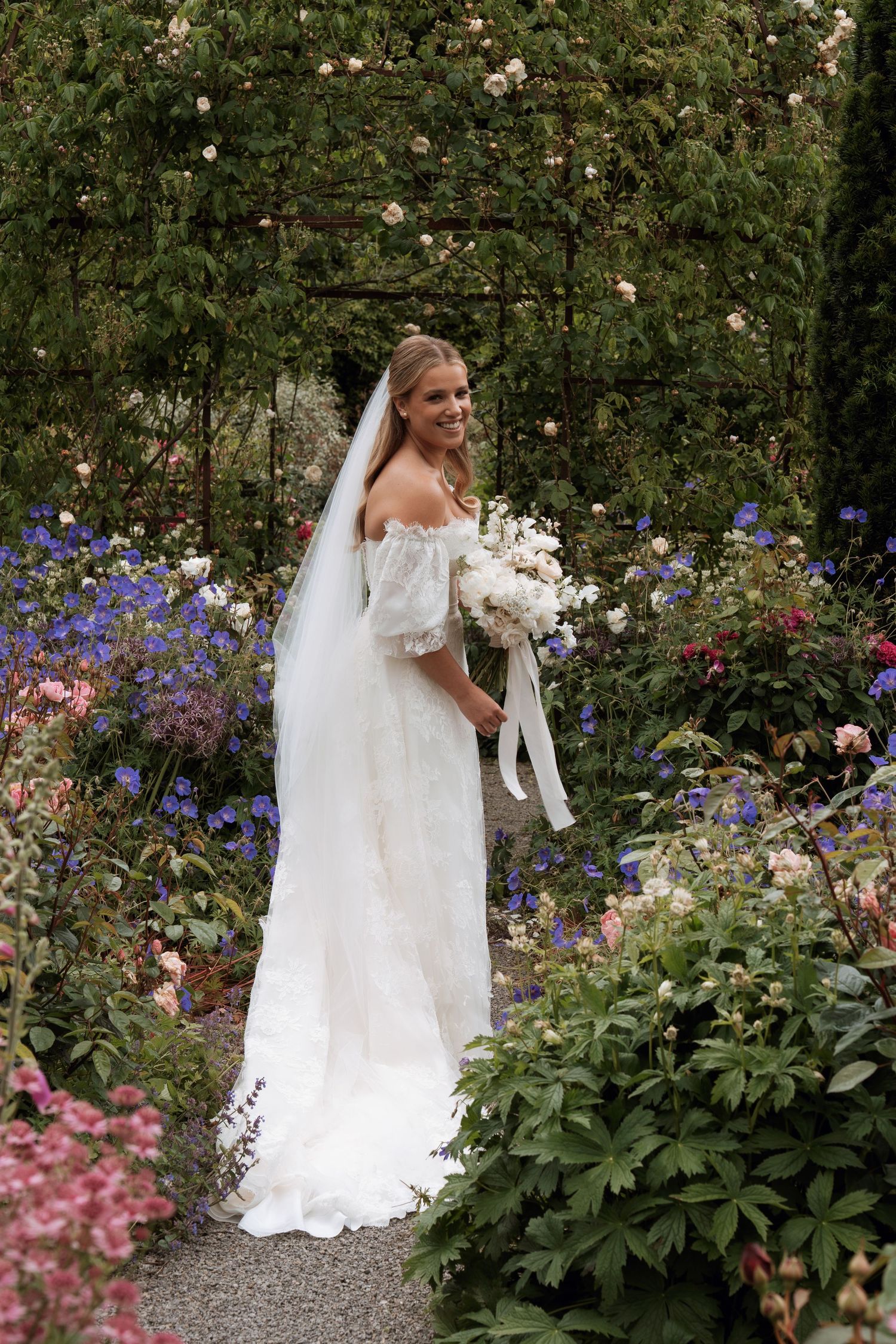 A bride in a flowing white wedding gown stands gracefully among colorful garden flowers and greenery.
