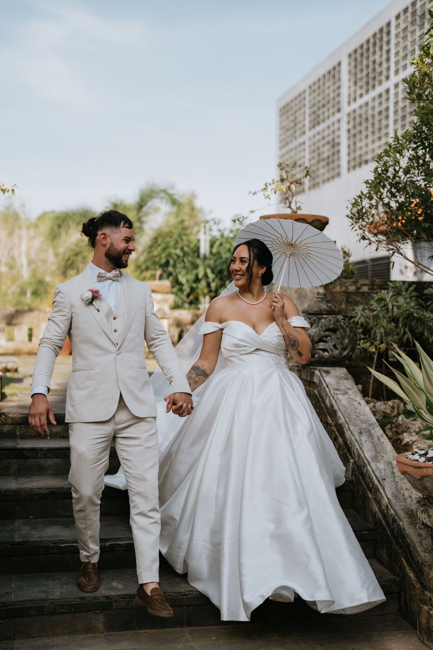 A couple walks down garden steps, bride holding parasol in elegant off-shoulder gown and groom in light suit.