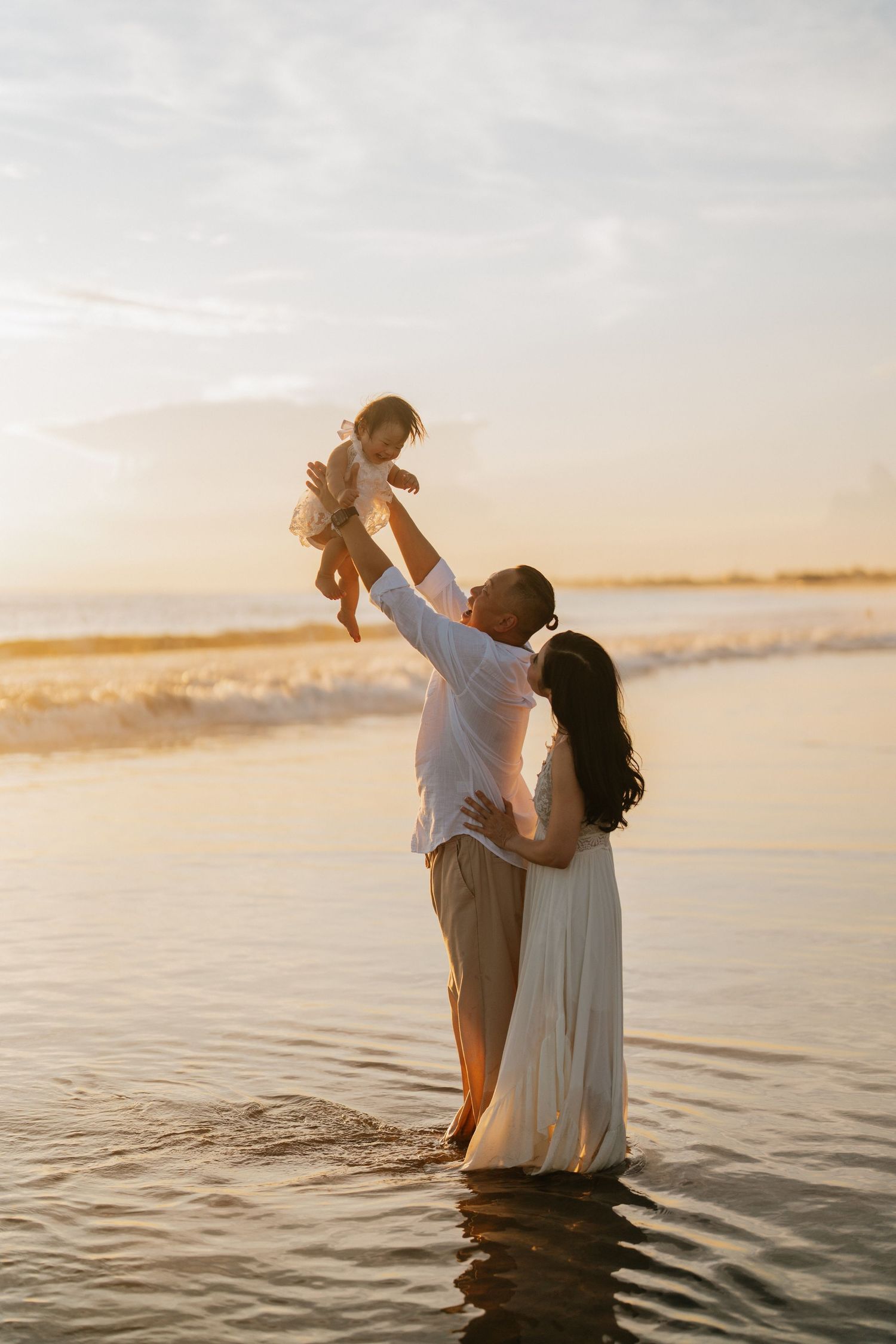Parent lifting child against golden sunset sky at beach shoreline.