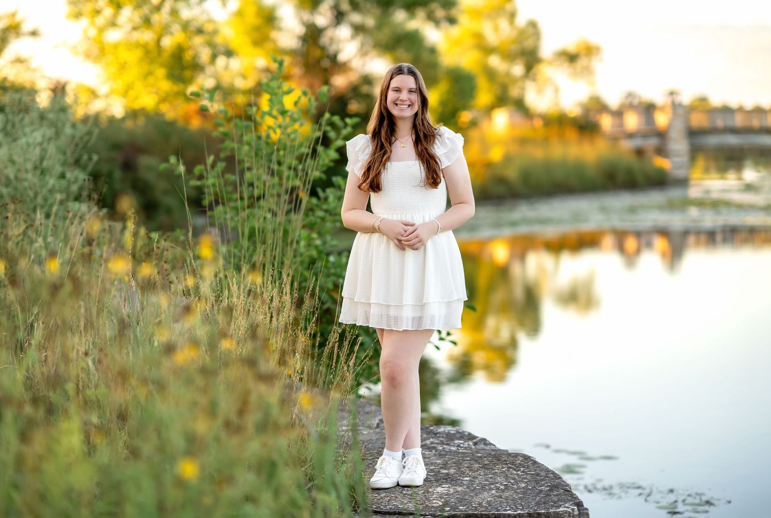 Woman in white dress poses for golden hour sunset outdoor photo session near tranquil lake surrounded by autumn foliage.