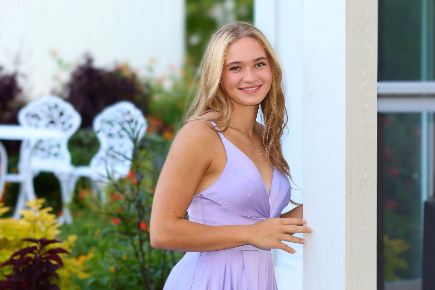 High school senior girl wearing a light purple gown leaning against a doorway in a garden at sunset.