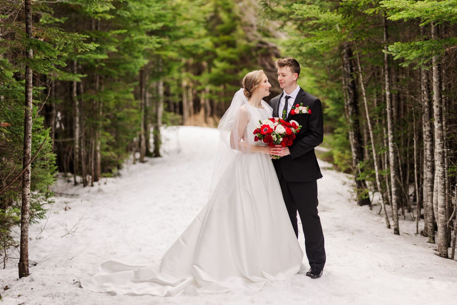 A couple in wedding attire embrace on a snowy forest path lined with evergreen trees.