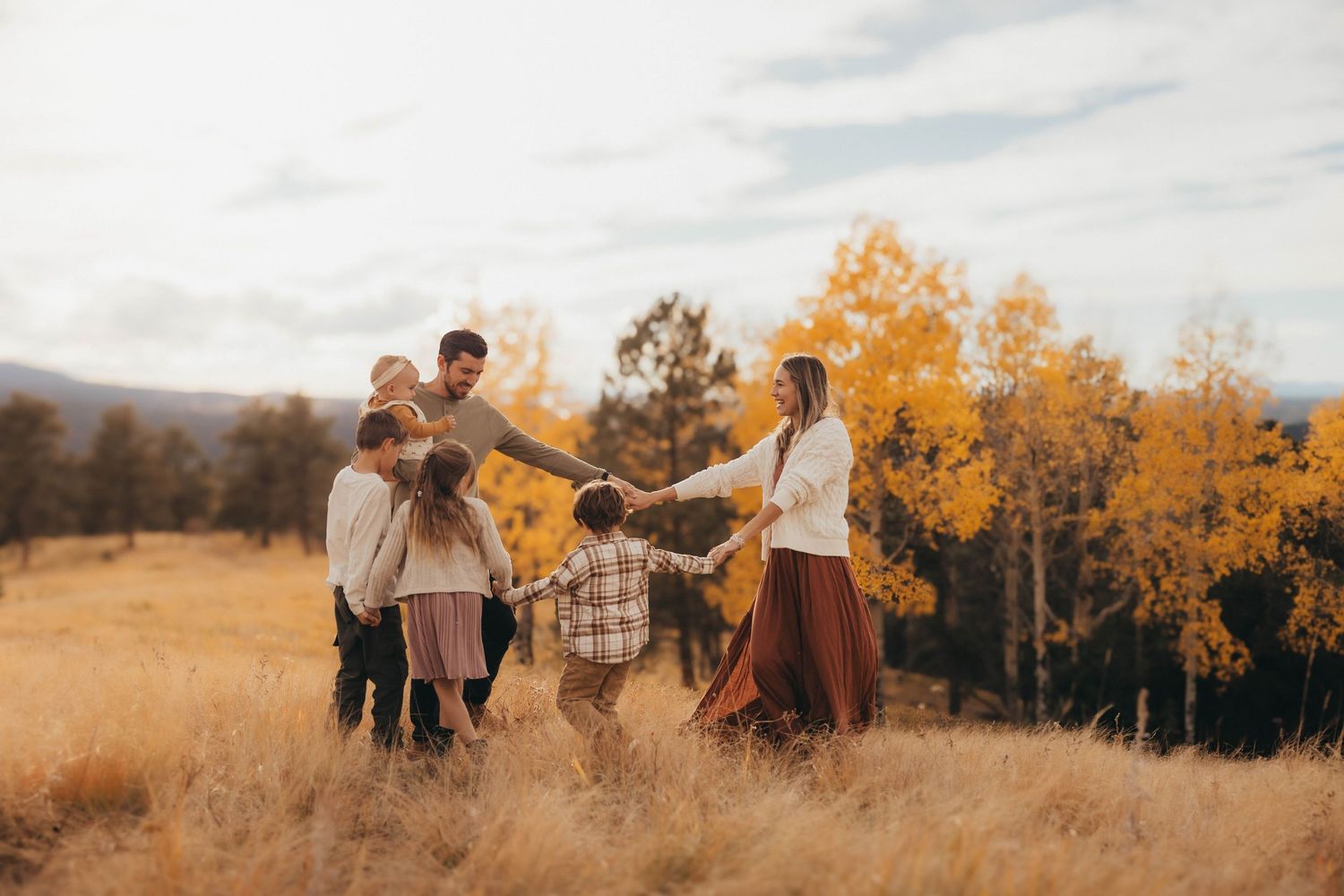Family holding hands in a circle in an autumn field with golden trees in the background.