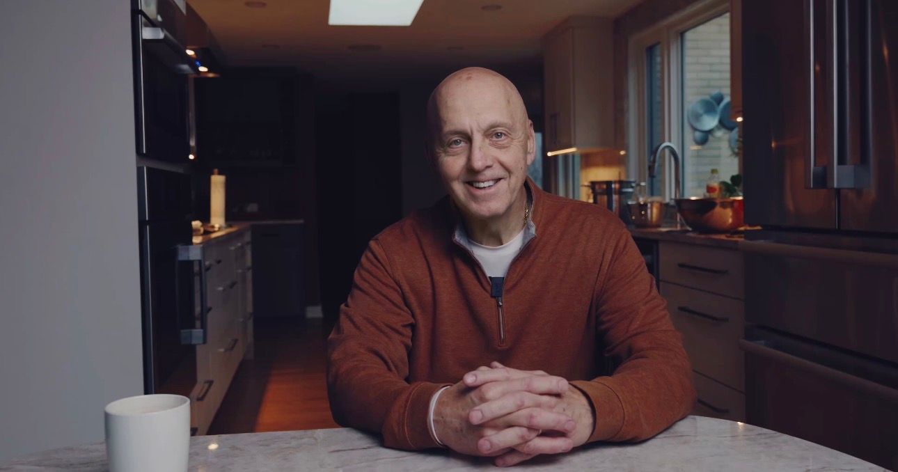 A person wearing a brown sweater sits at a white table with their hands clasped in a bright indoor setting.