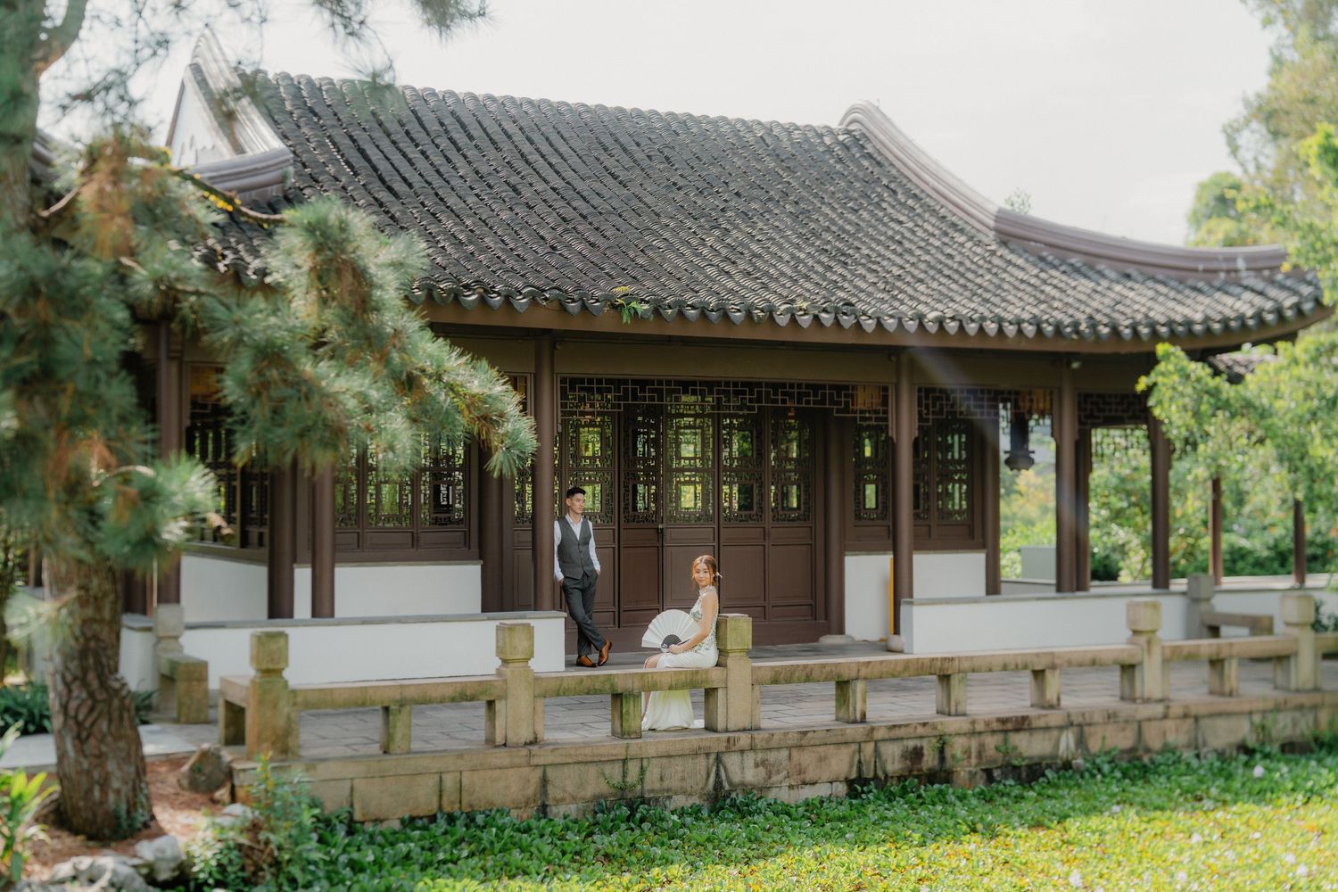 A winding pathway leads to a traditional Chinese garden pavilion with a stone bridge and weeping willow trees.
