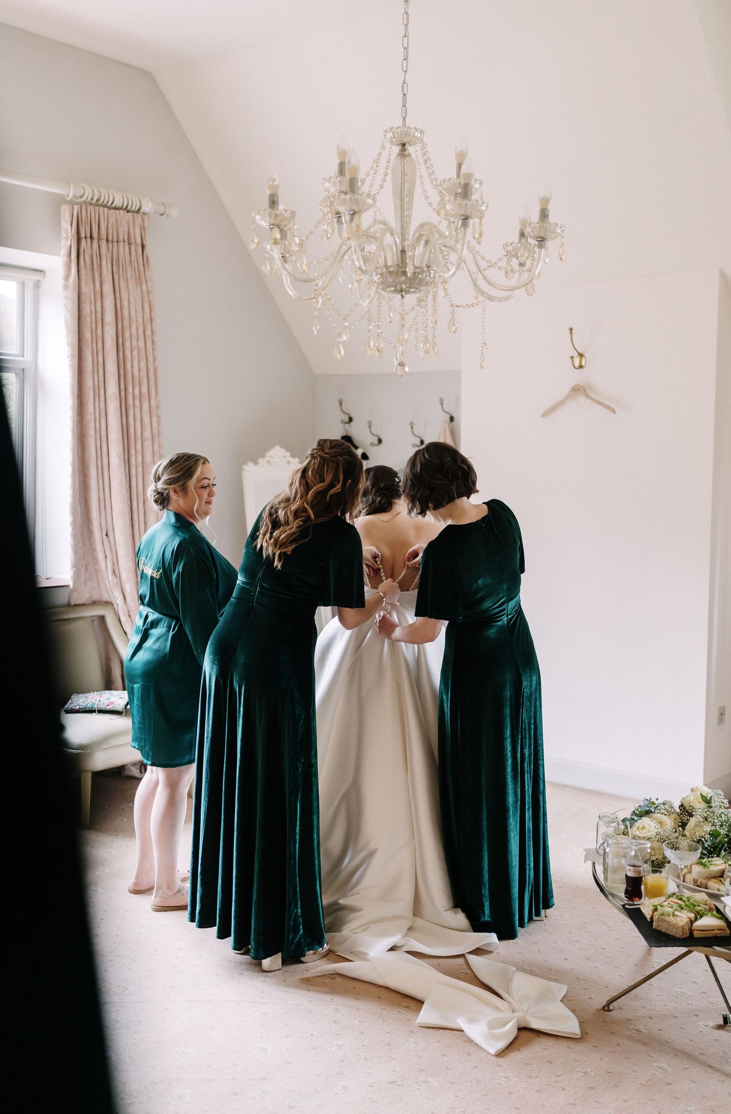 A group in emerald green velvet dresses helps with wedding preparations in an elegant white room with chandeliers.