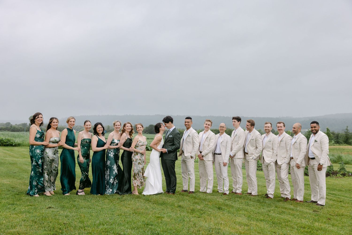 Wedding party in green dresses and light tan suits poses together on scenic grassy hillside overlooking ocean views.