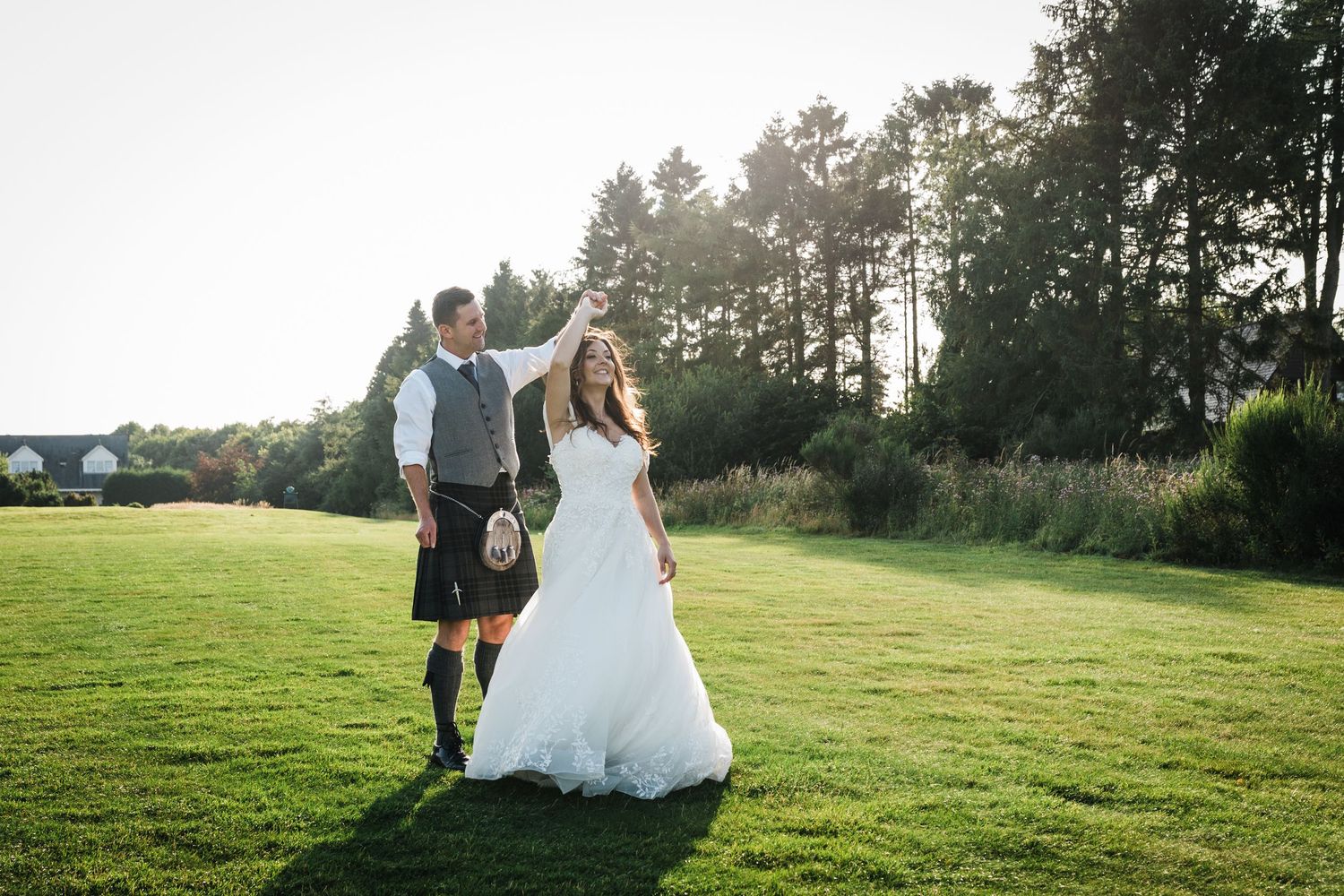 Groom in kilt and bride dancing on Forbes of Kingennie golf course.  Full body colour shot with trees in background at golden hour
