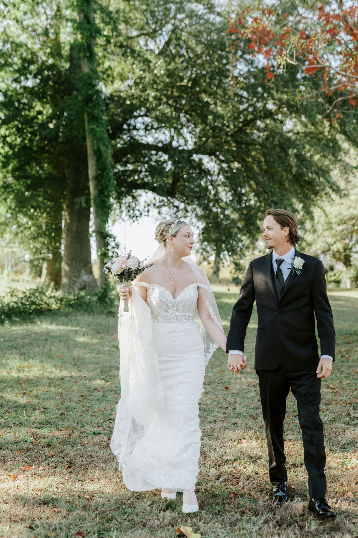 Bride and groom walk hand in hand through a sunlit garden with lush green trees and autumn leaves.