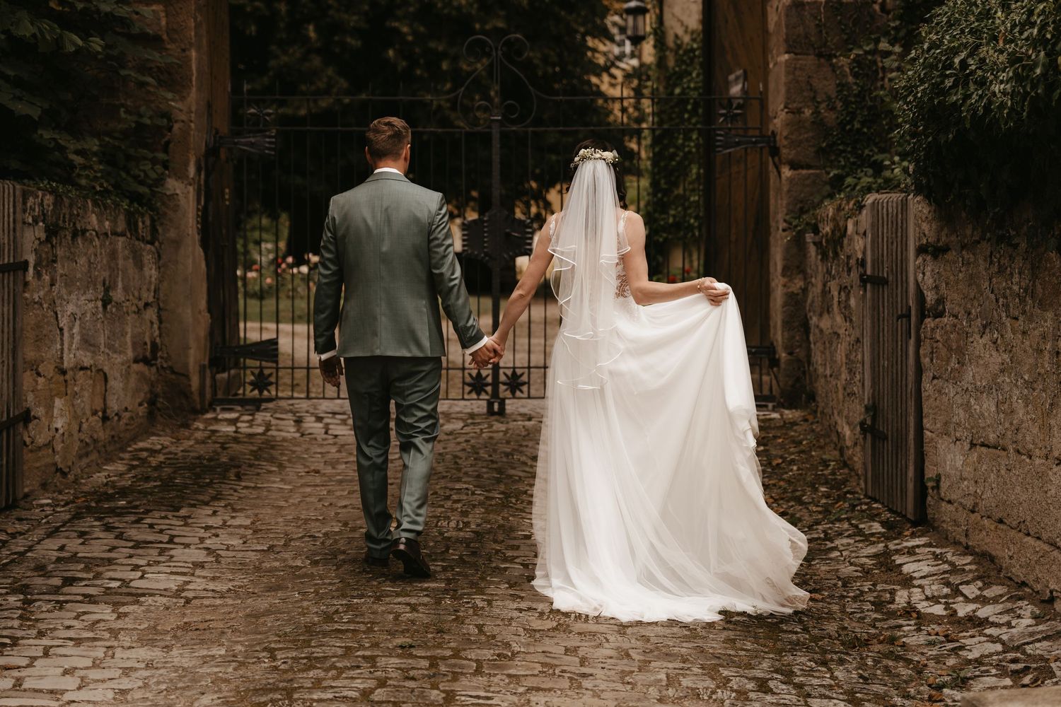 A couple in wedding attire walks through a stone archway on a cobblestone path.