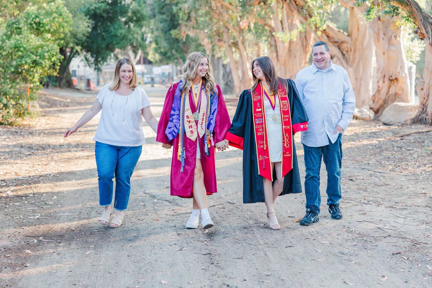 Group of graduates in red regalia and their companions walking together on a dirt path under shady trees.