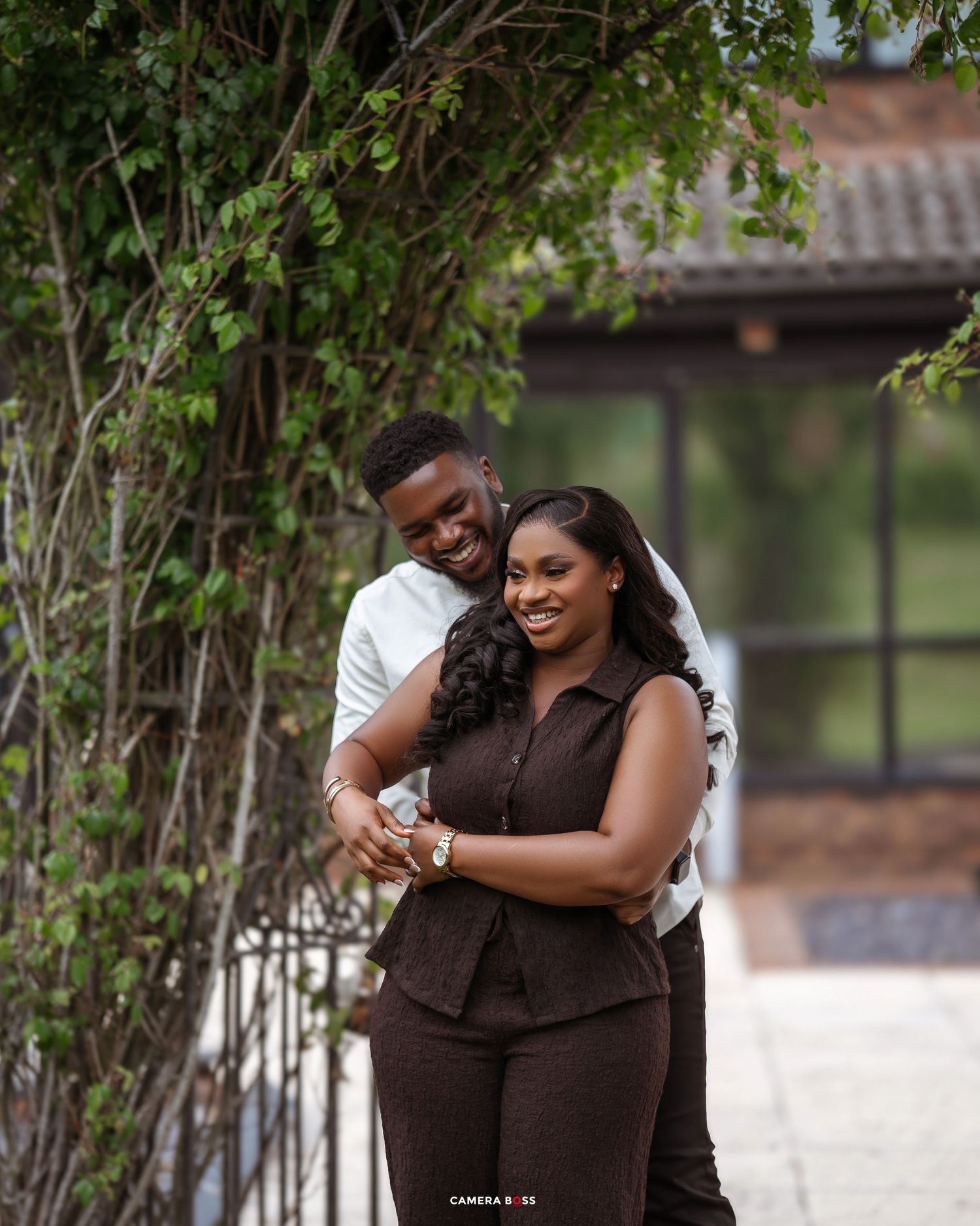 A couple shares a romantic moment outdoors while embracing near lush greenery and trees.