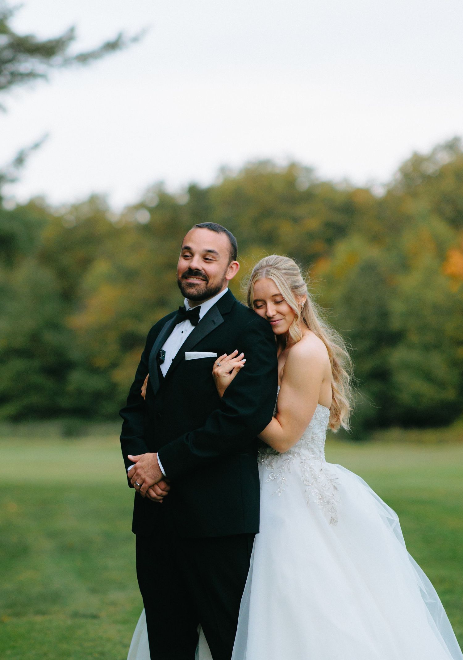 A happy couple shares a playful first look moment in a beautiful outdoor setting with autumn trees in the background.