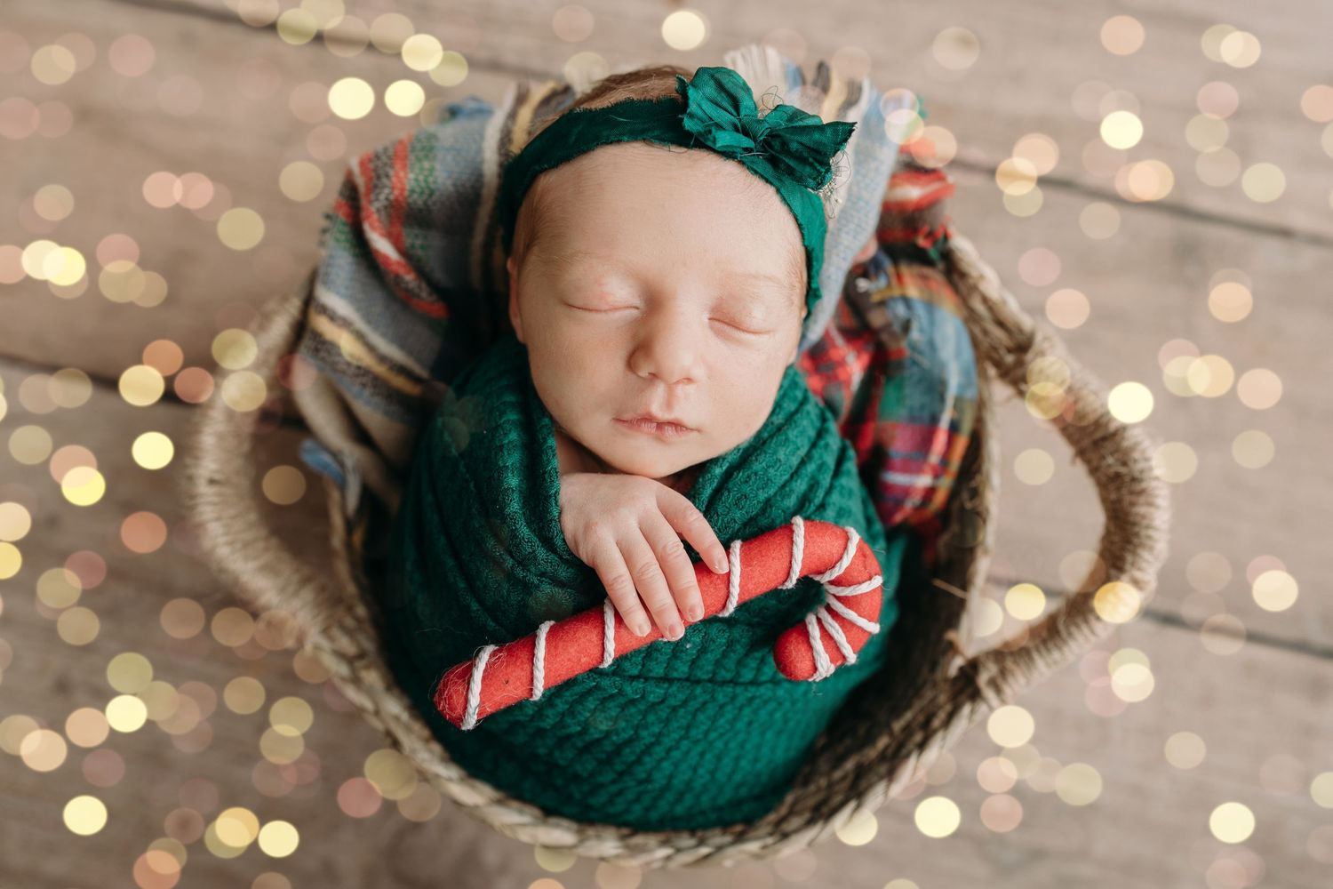 Sleeping newborn swaddled in emerald green wrap and headband lying in basket with twinkling holiday lights in background.