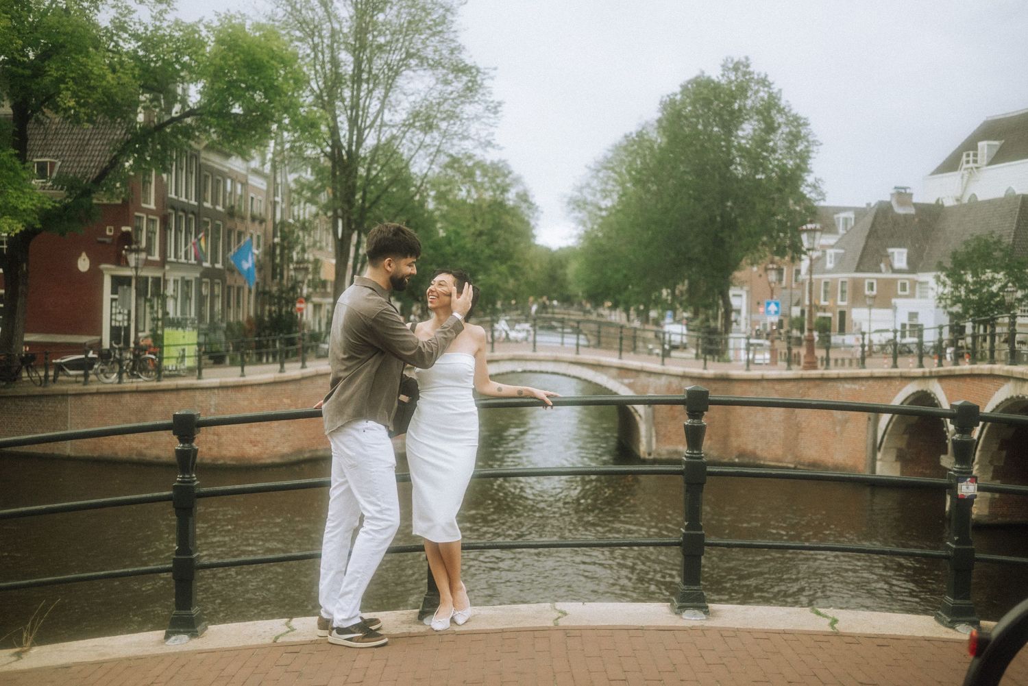 Couple embracing on a canal bridge in Amsterdam, with historic brick buildings and tree-lined waterways in the background.