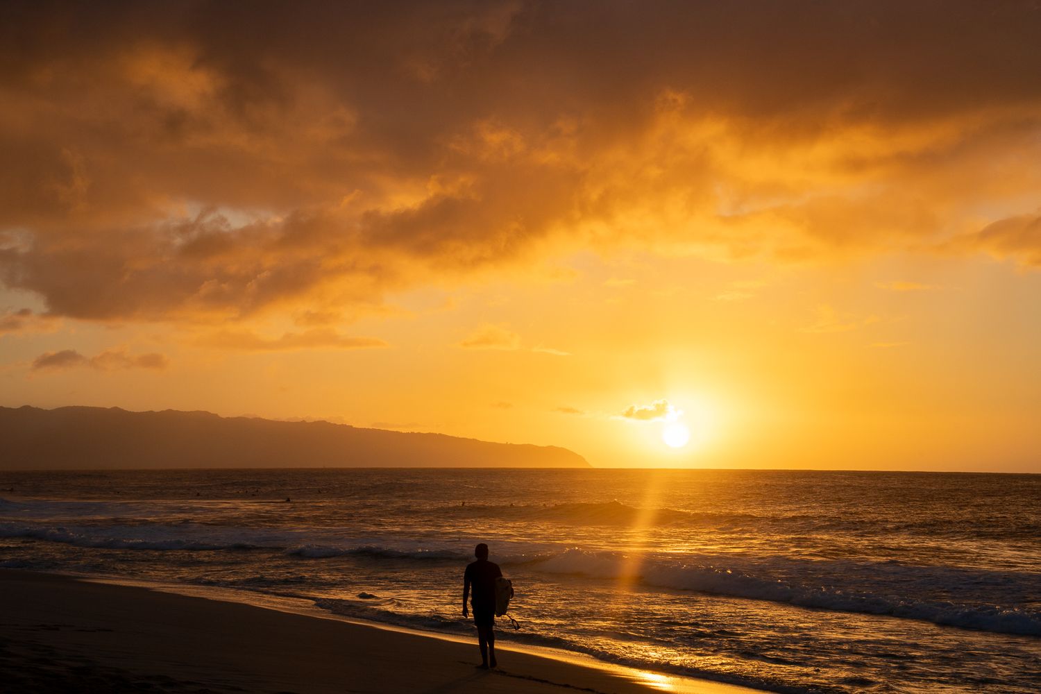 A silhouetted person walks along a beach at sunset with dramatic orange clouds reflecting on the ocean waves.